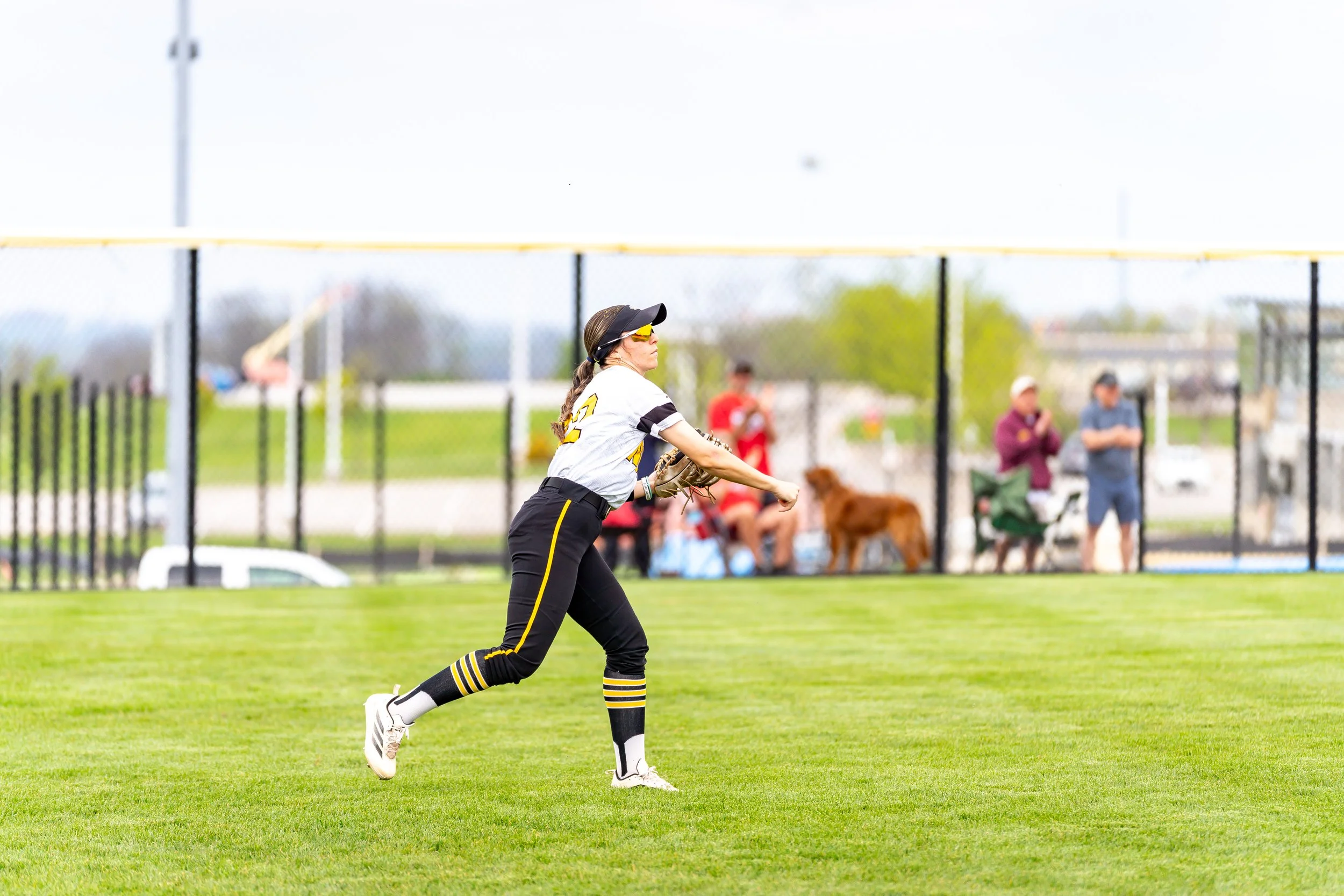 A woman playing softball on a field, wearing black athletic pants, a white jersey with black and yellow accents, black and yellow striped socks, white cleats, a black visor, and sunglasses. She is in the middle of a throwing motion. In the background