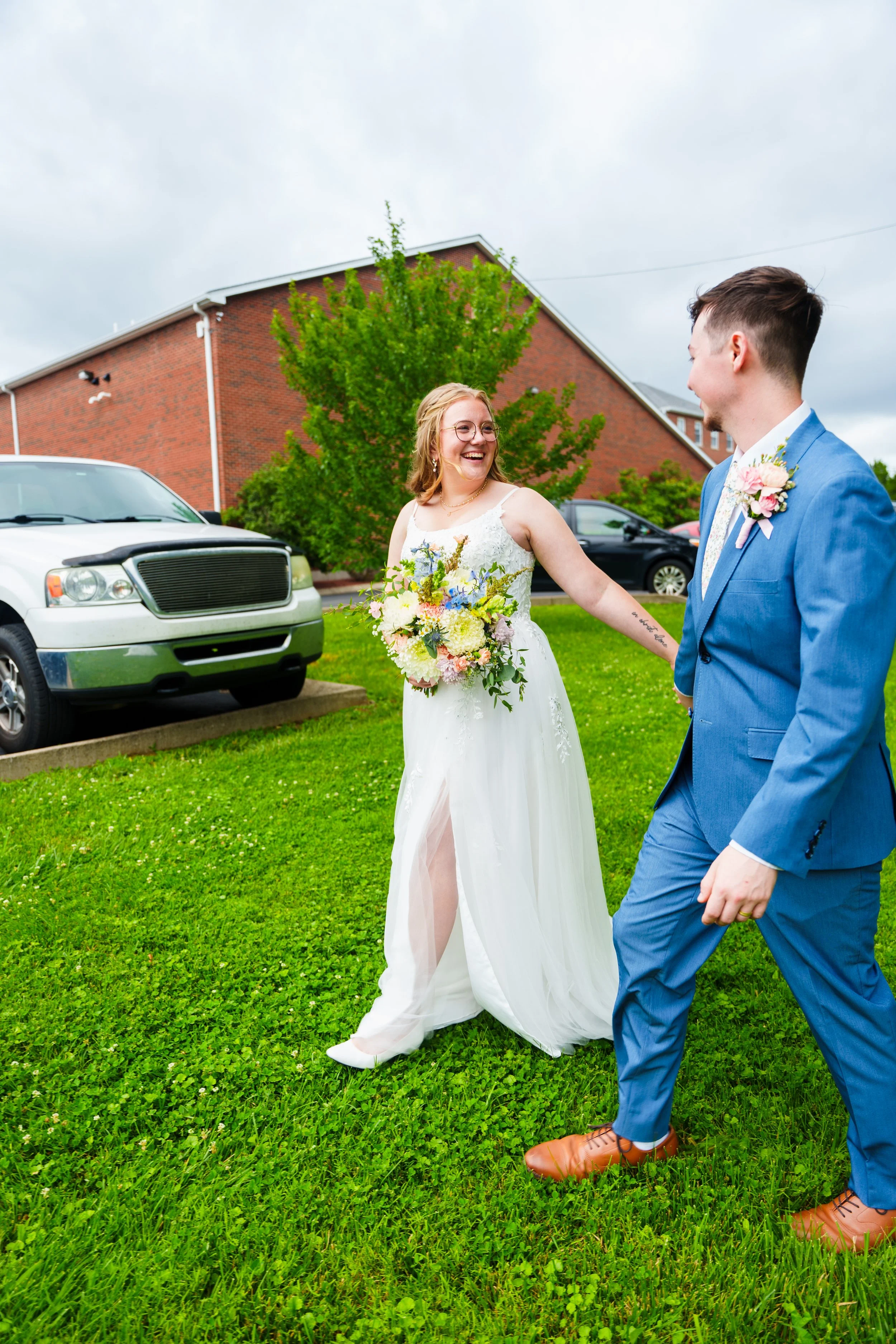 A smiling woman in a white wedding dress holding a bouquet of flowers walking outdoors with a man in a blue suit in front of a brick building and a parking lot on a cloudy day.