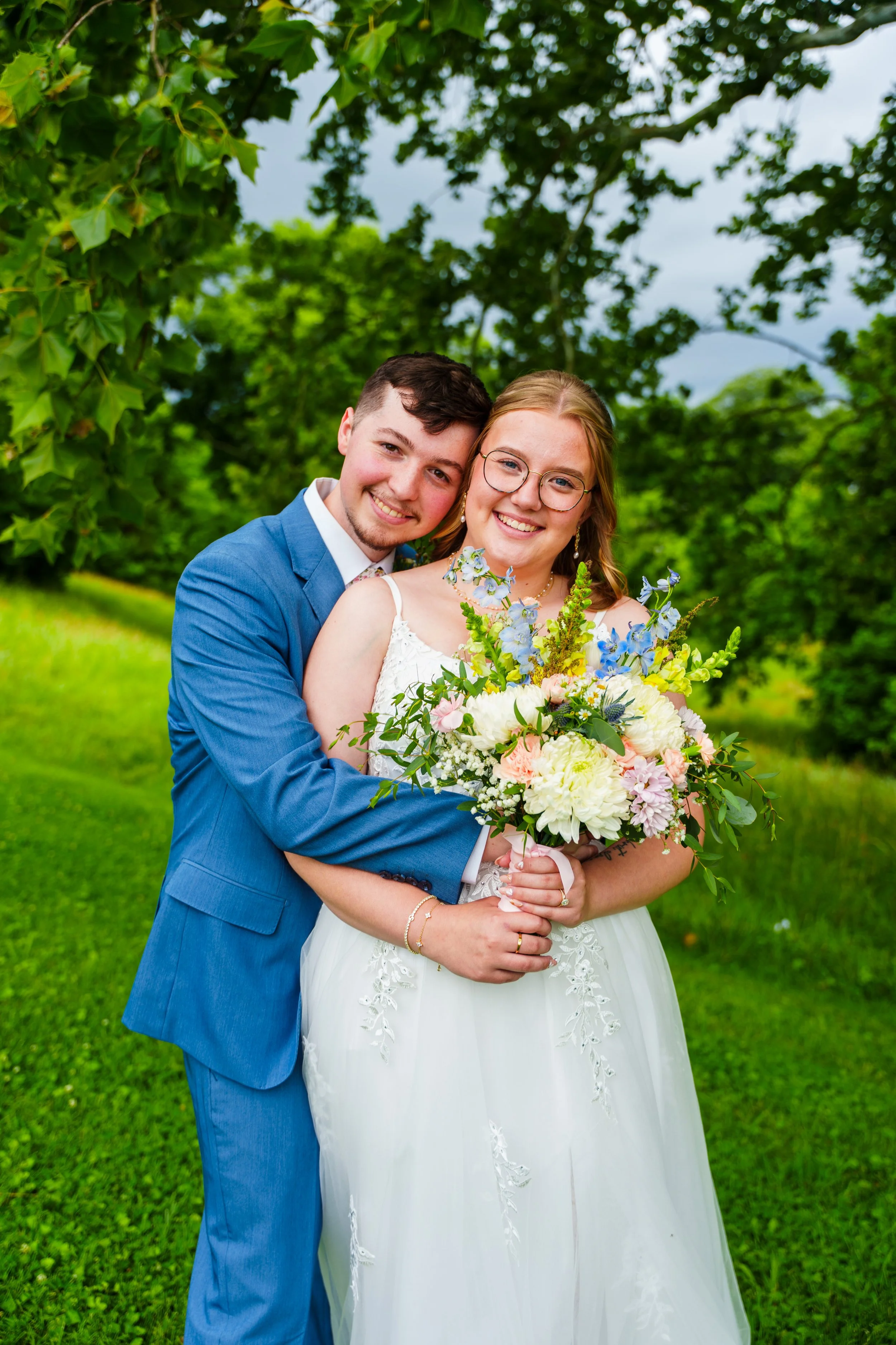 A newlywed couple standing outdoors on a lush green lawn, smiling and holding a large bouquet of flowers, surrounded by trees.