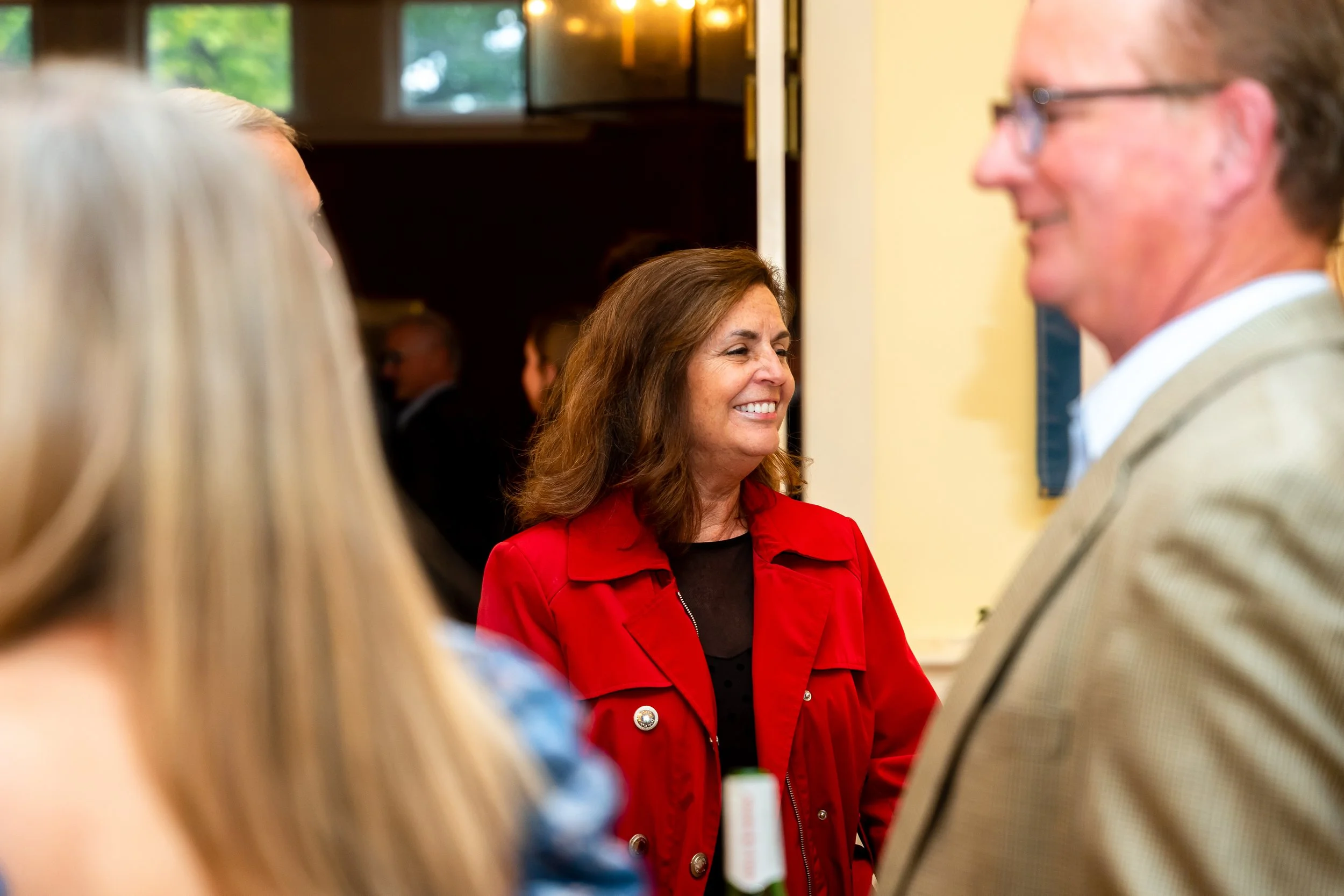 A woman with brown hair smiling while talking to a man wearing glasses. The woman is wearing a red jacket and is in focus, while the man is slightly blurred.