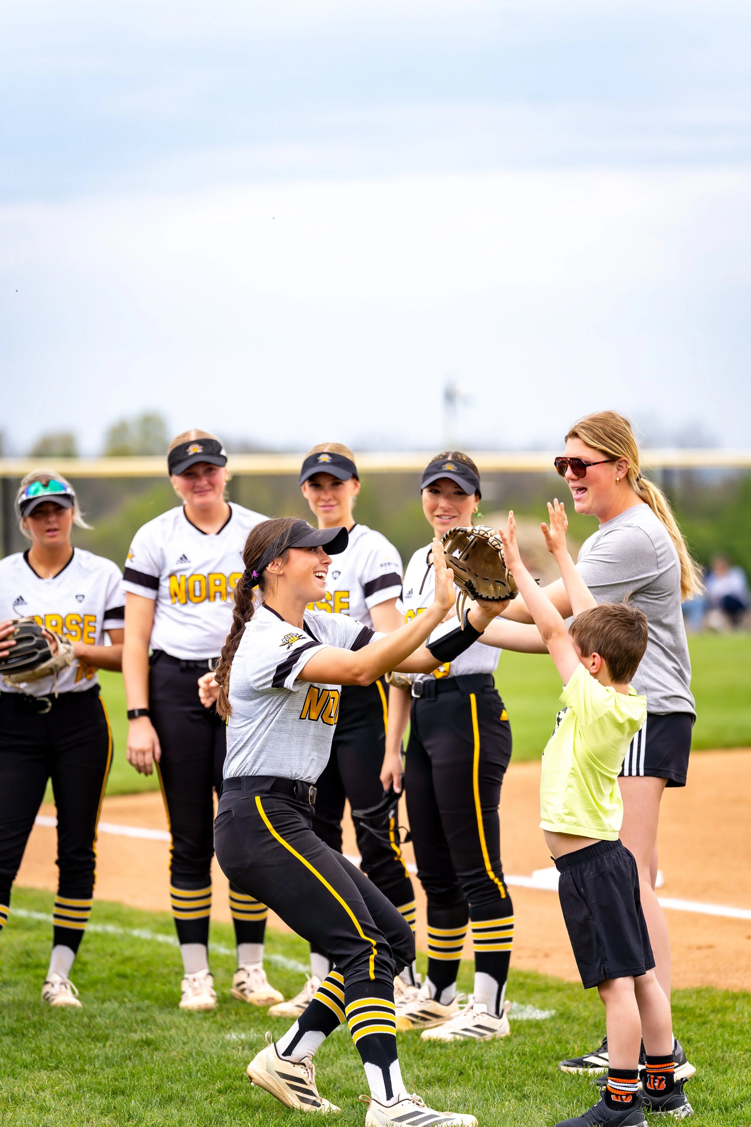 A group of young female softball players in black and yellow uniforms standing on a baseball field, smiling and celebrating. An adult woman is giving high-fives to a young boy in a neon green shirt. The background shows a cloudy sky and a baseball fi