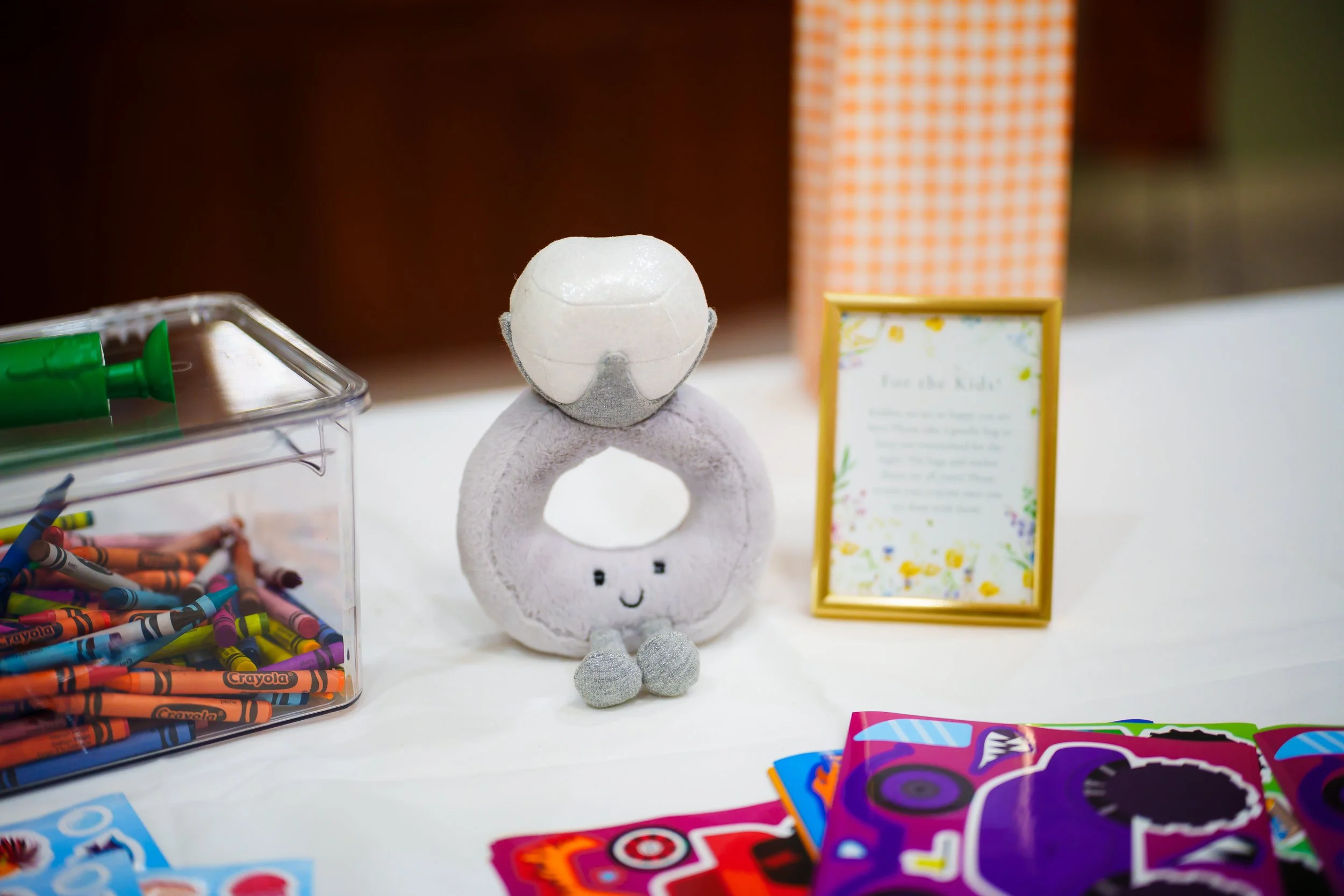 Table with various colorful arts and crafts supplies including crayons, markers, and stickers, with a plush toy shaped like a toilet with a smiling face at the center.
