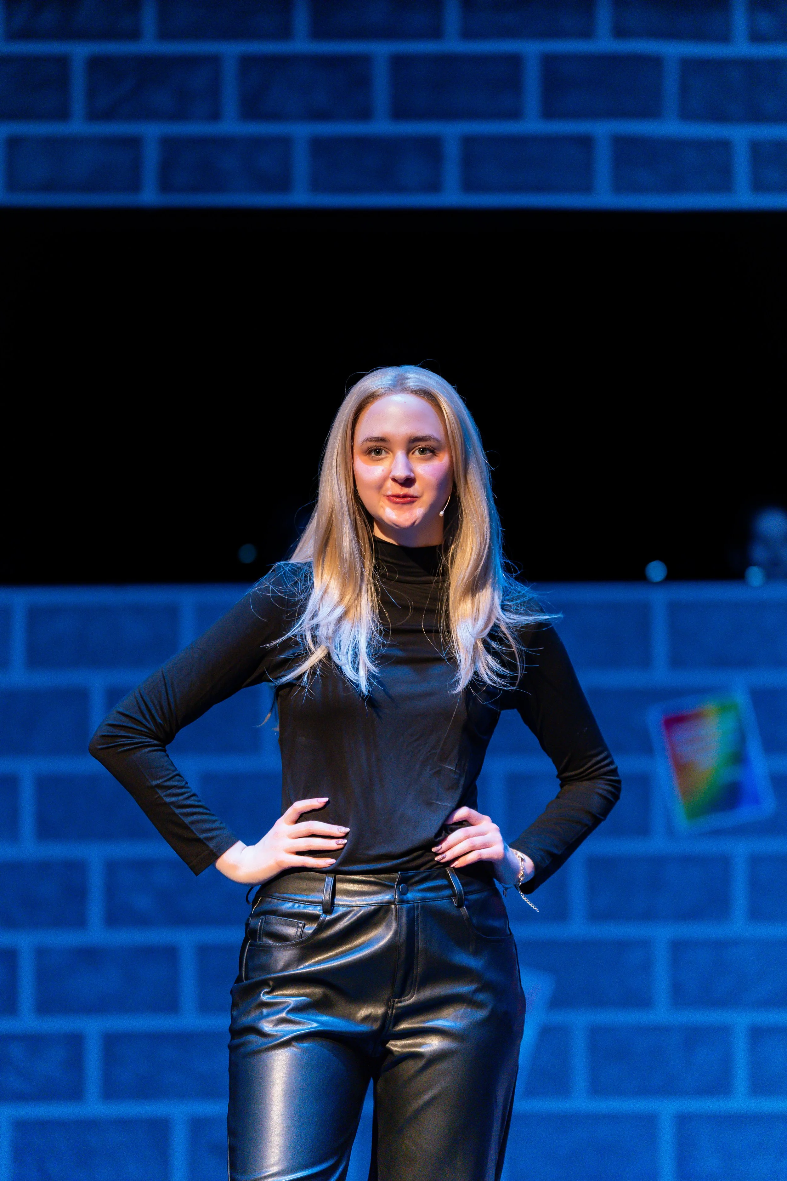 Young woman with long blonde hair wearing a black long sleeve top and leather pants, standing with hands on hips in front of a blue brick wall.