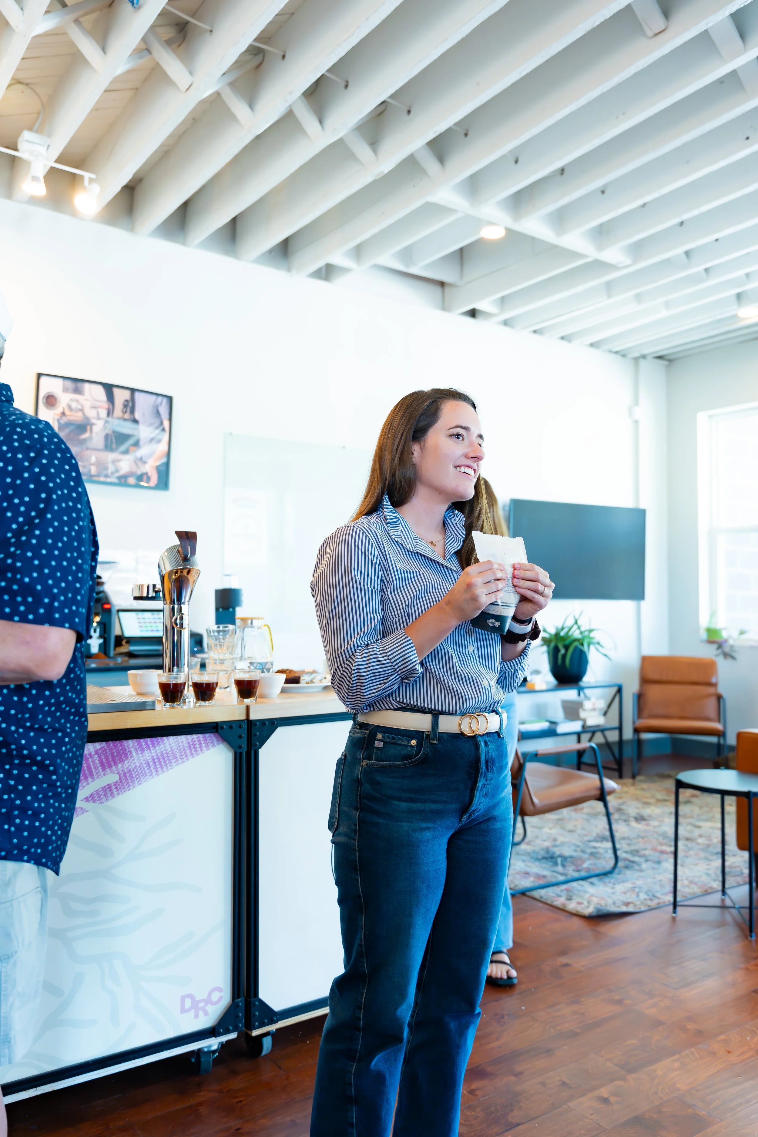 A woman with long brown hair, wearing a blue and white striped shirt and blue jeans, is smiling and holding a paper cup in a modern, well-lit room with a wooden floor, large window, and chairs
