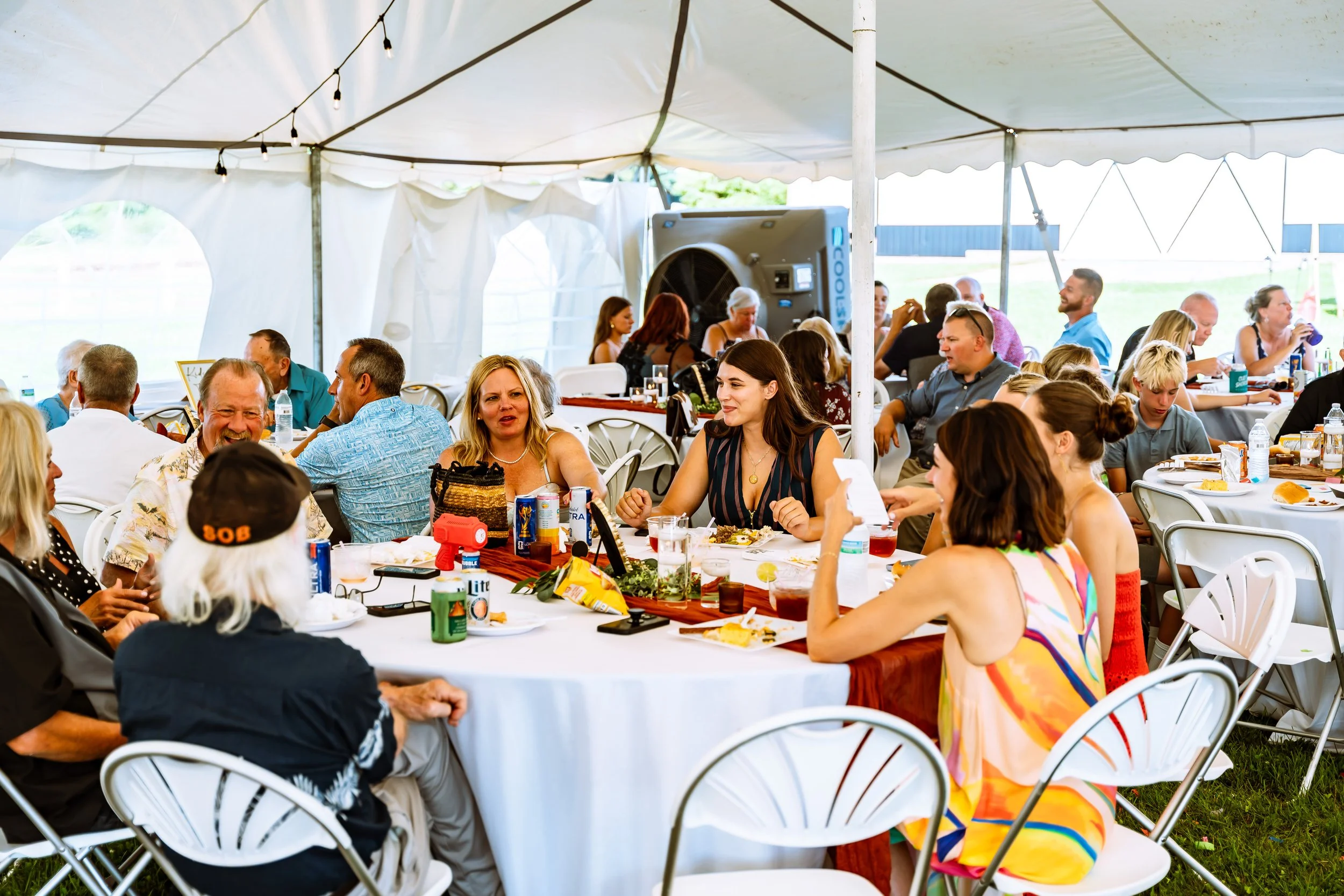 People gathered at a long outdoor picnic table under a tent, enjoying food and conversation.