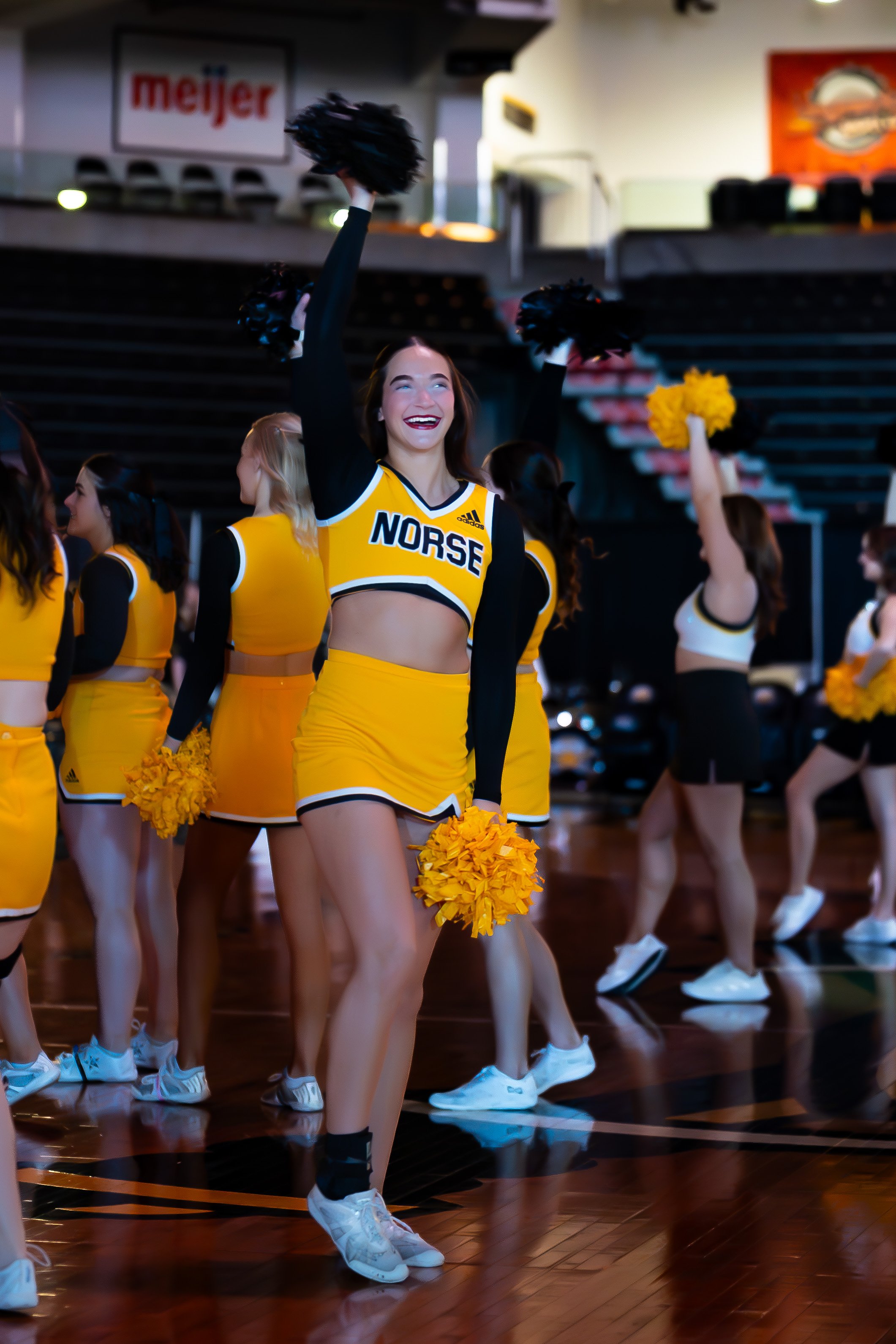 Cheerleader in yellow and black uniform with 'Norse' written on it, holding yellow pom-poms and smiling during a cheerleading routine in a gymnasium.