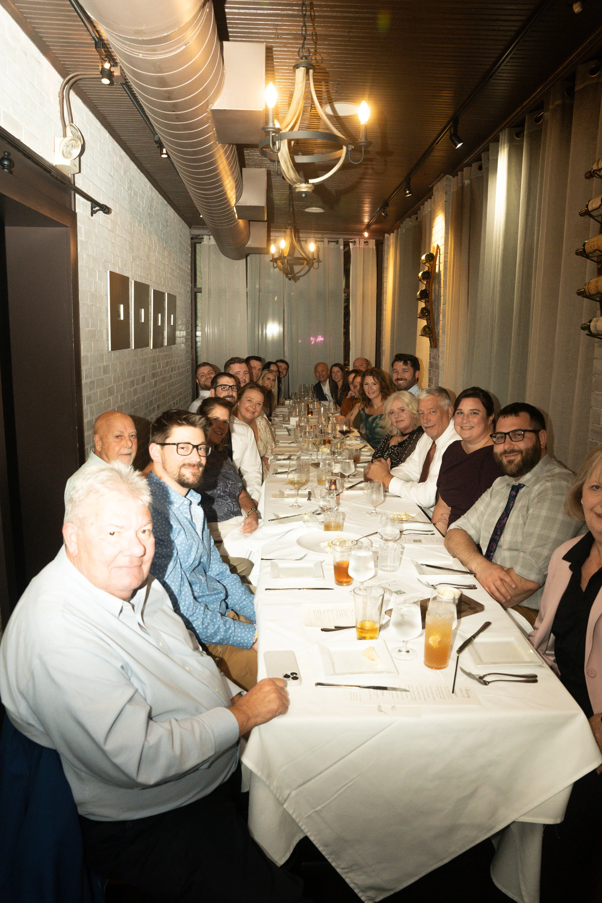 A large group of people sitting around a long dining table at a restaurant, smiling and posing for the photo. The table is set with drinks, menus, and utensils, with a brick wall and hanging lights in the background.