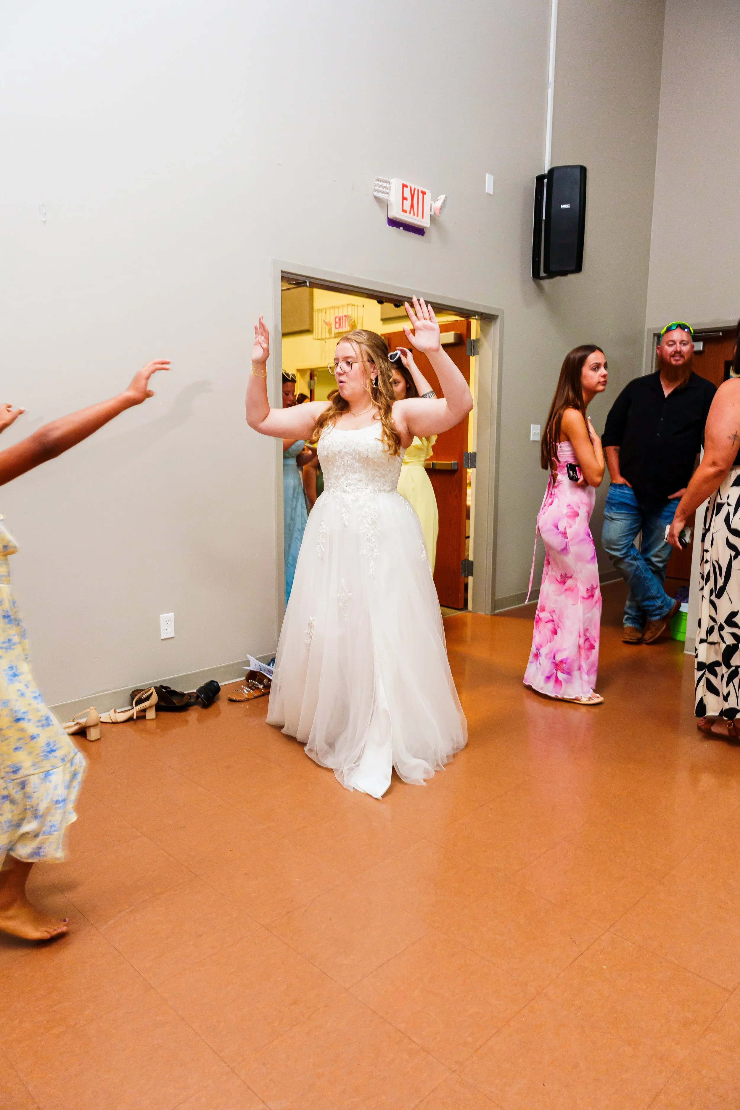 A woman in a white wedding dress raising her hands while standing on a dance floor at a party or event. Several people are present in the background, some in colorful, casual attire, suggesting a celebration or gathering.