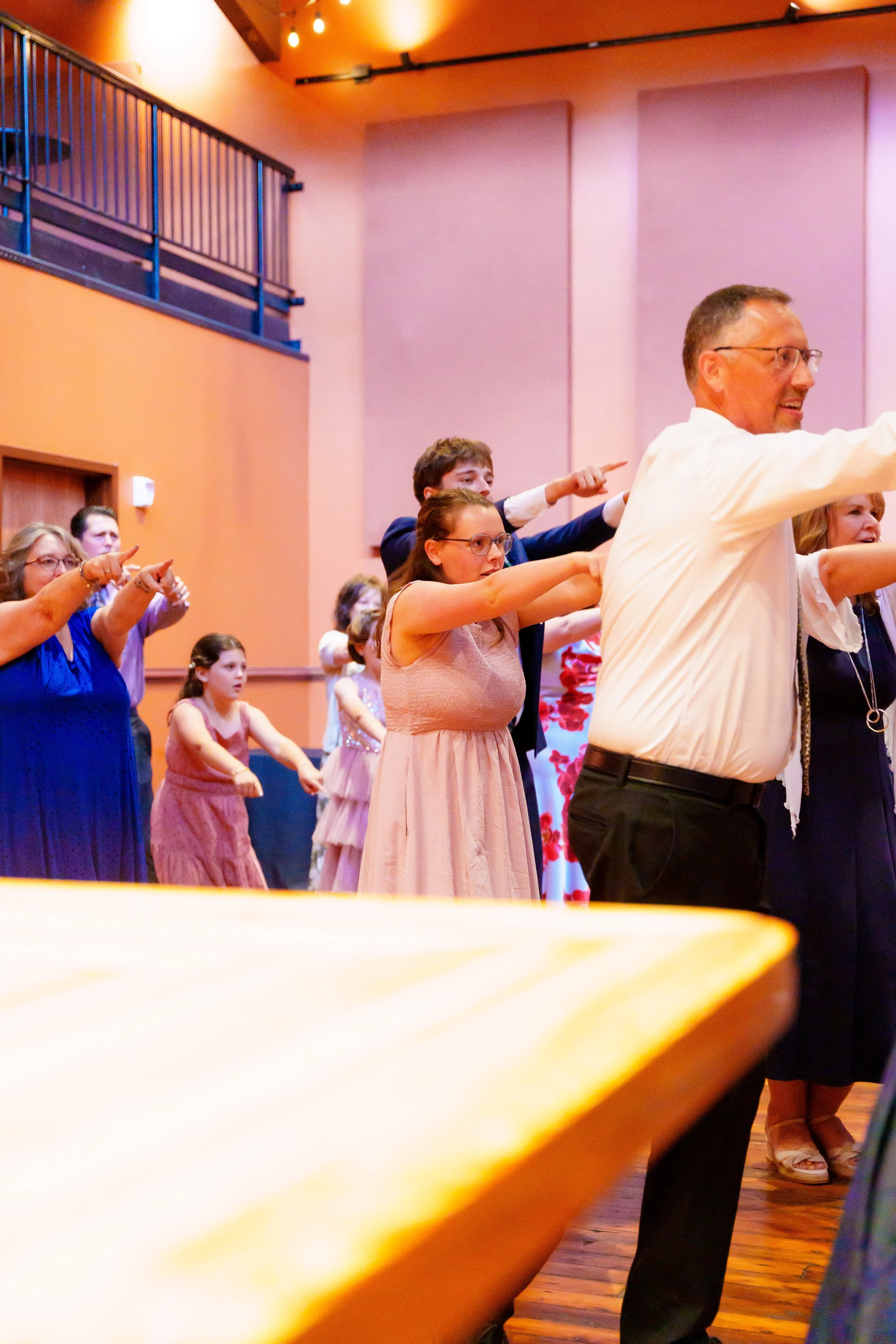A group of people dancing in a room with pink walls, some children and adults, all pointing forward with one arm extended.