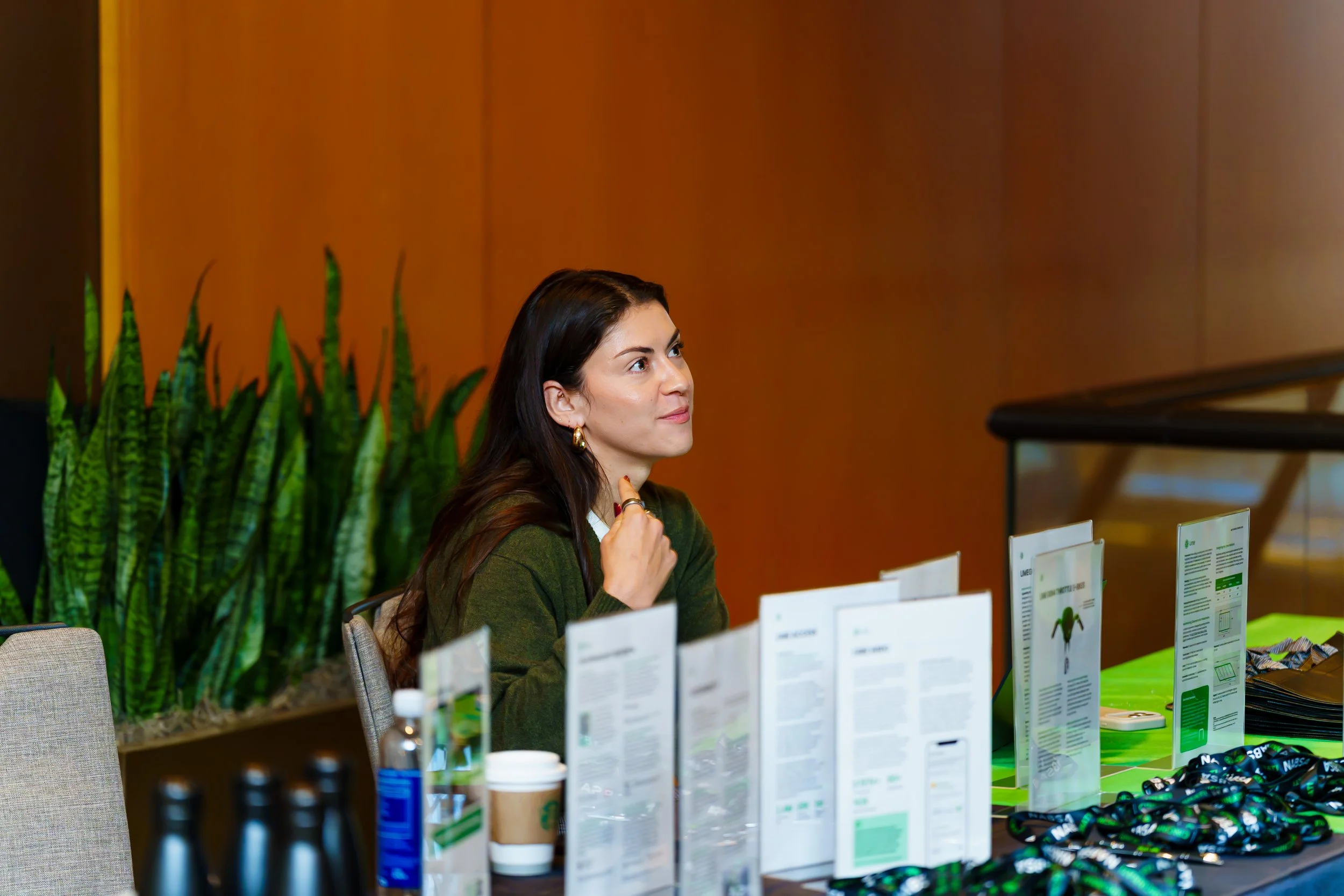 A woman with dark hair, wearing a green sweater and gold hoop earrings, seated at a table with promotional materials, brochures, and items like bottled water and coffee cups, in an indoor setting with warm-colored walls and a green plant in the background.