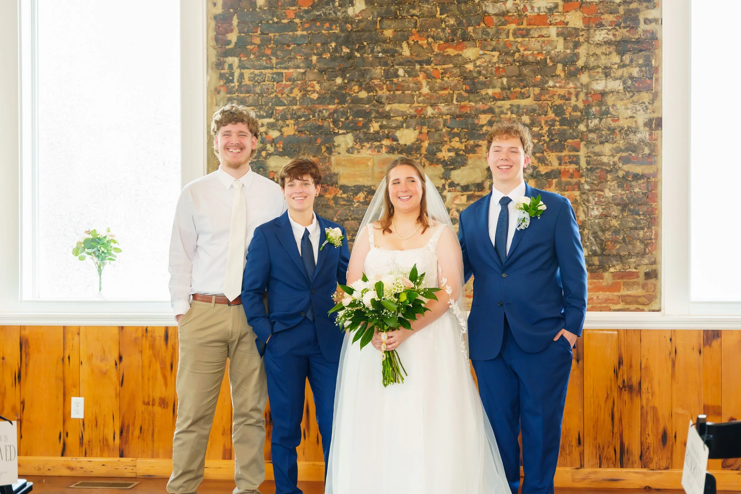 Group of four people at a wedding, two men and two women, standing indoors against a brick wall. The bride is in the center holding a large bouquet of flowers, wearing a white wedding dress and veil. Two men are dressed in blue suits with flower bout