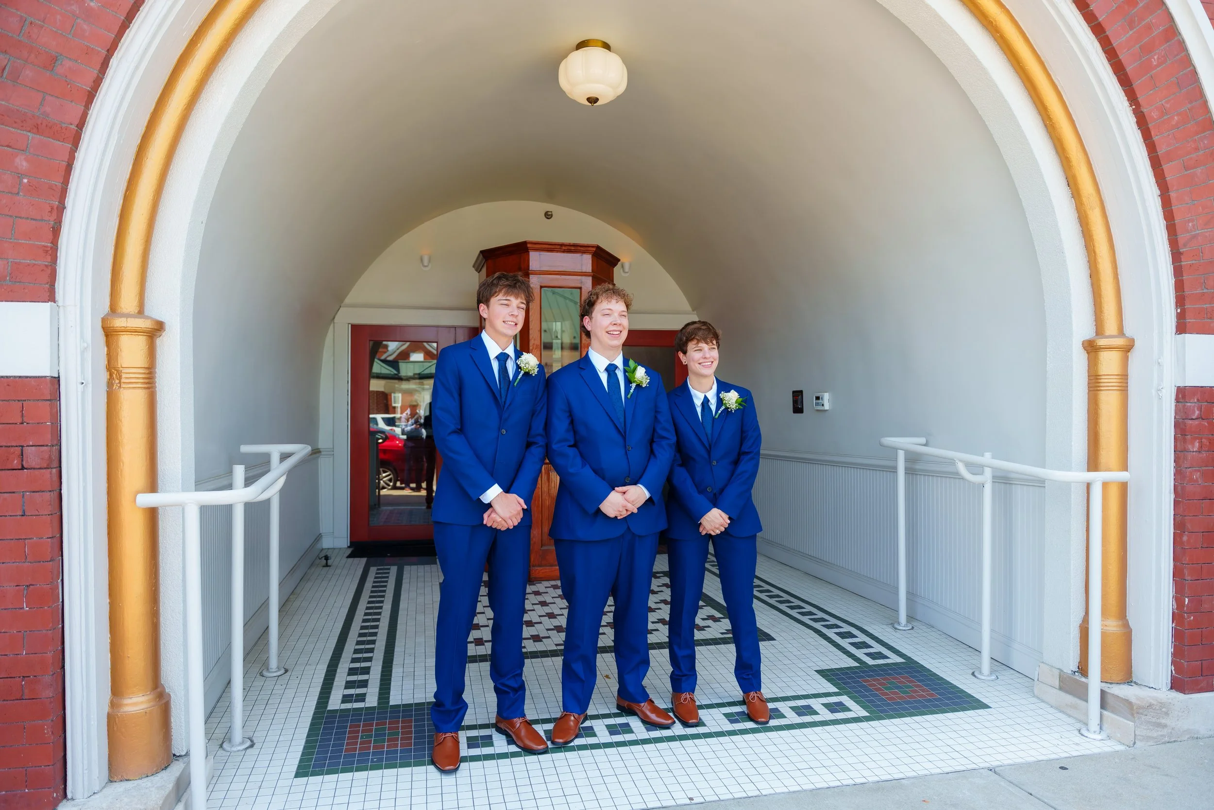 Three young men in blue suits and brown shoes standing at the entrance of a building with brick and arched doorway, smiling.