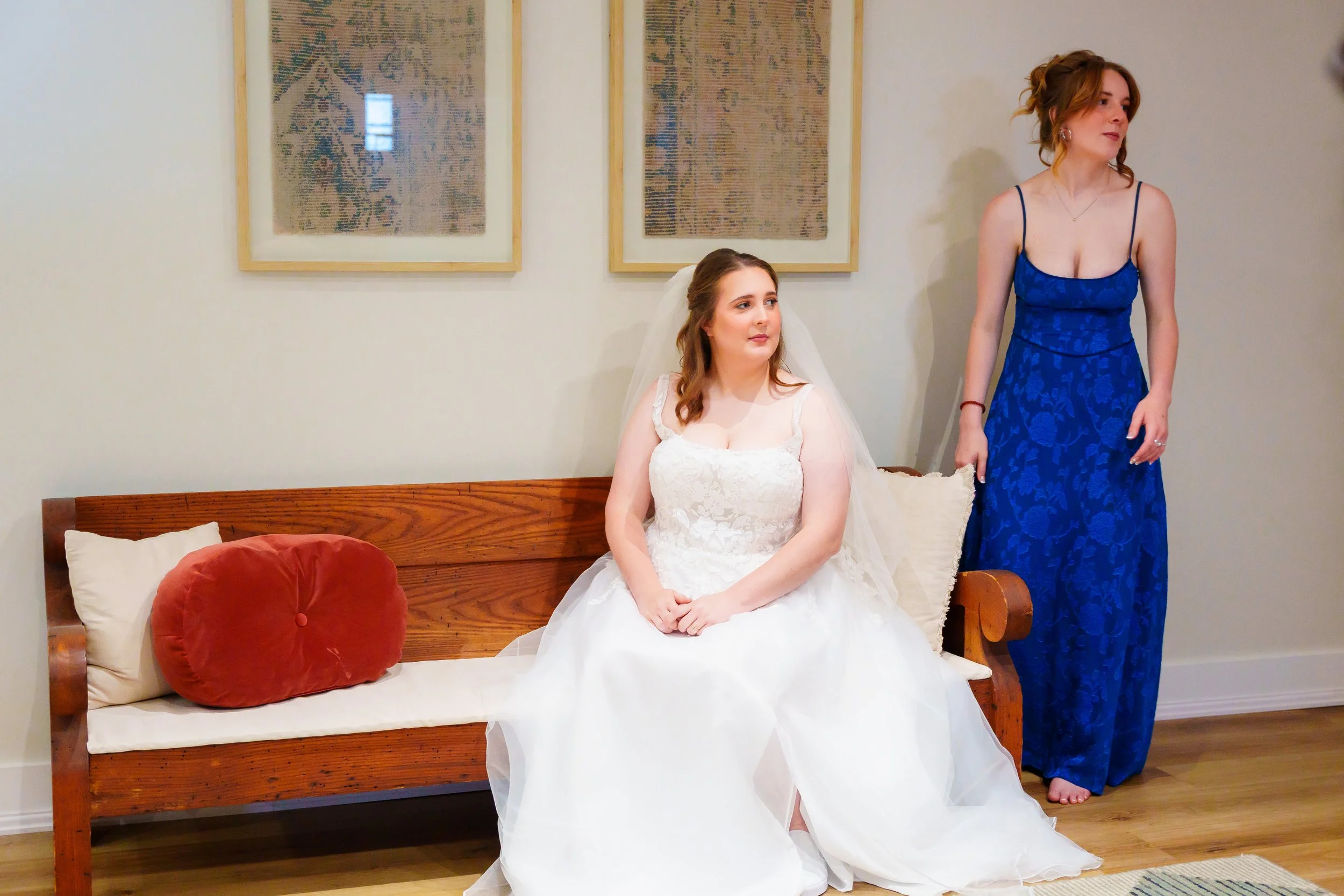 A bride in a white wedding dress sitting on a wooden bench with pillows, looking to her right, and a woman in a blue dress standing beside her, holding a cloth, in a room with framed artwork on the wall.