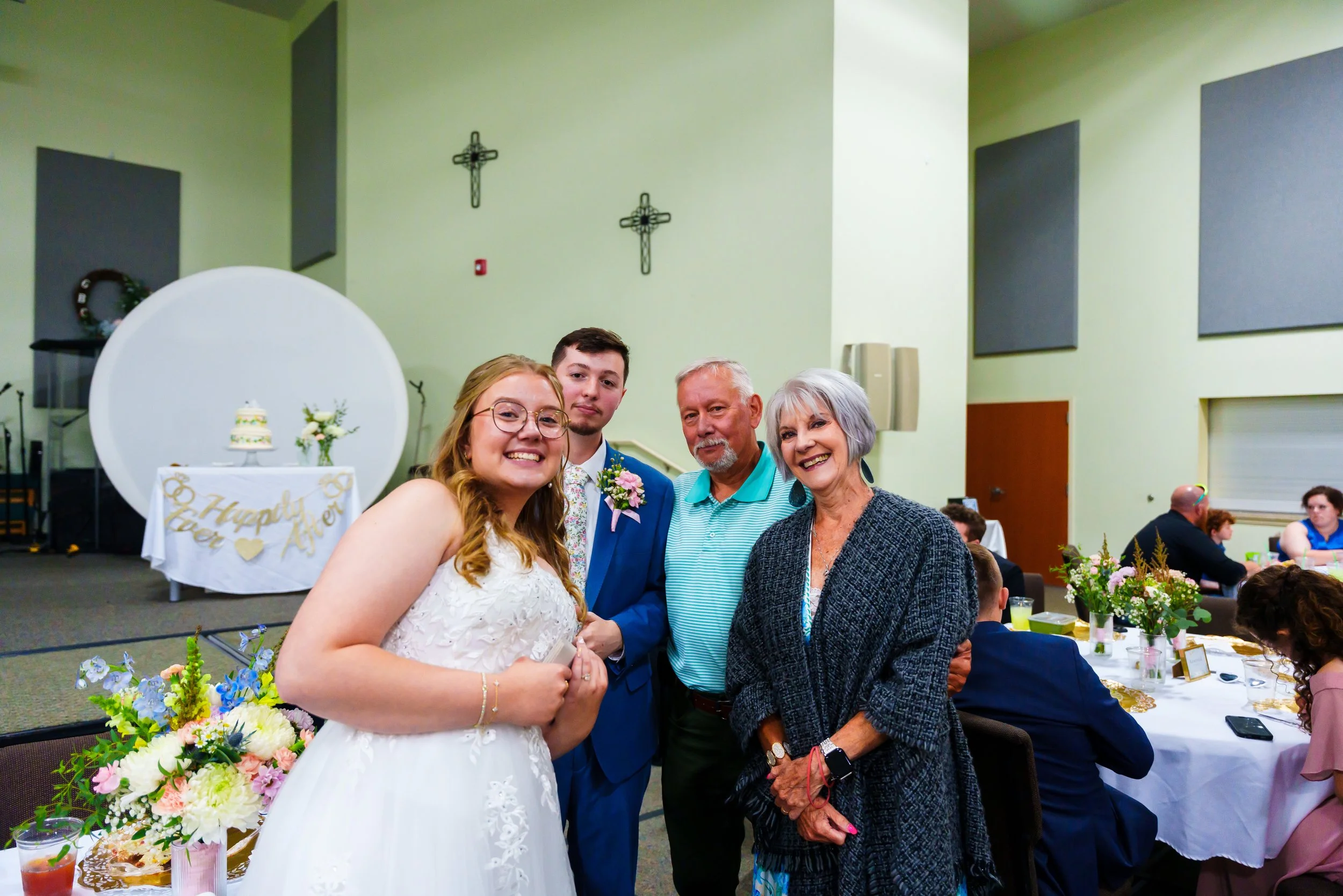 A joyful young woman in a white wedding dress standing next to three older adults, possibly her parents, at a wedding reception in a decorated hall with flowers, tables, and guests.