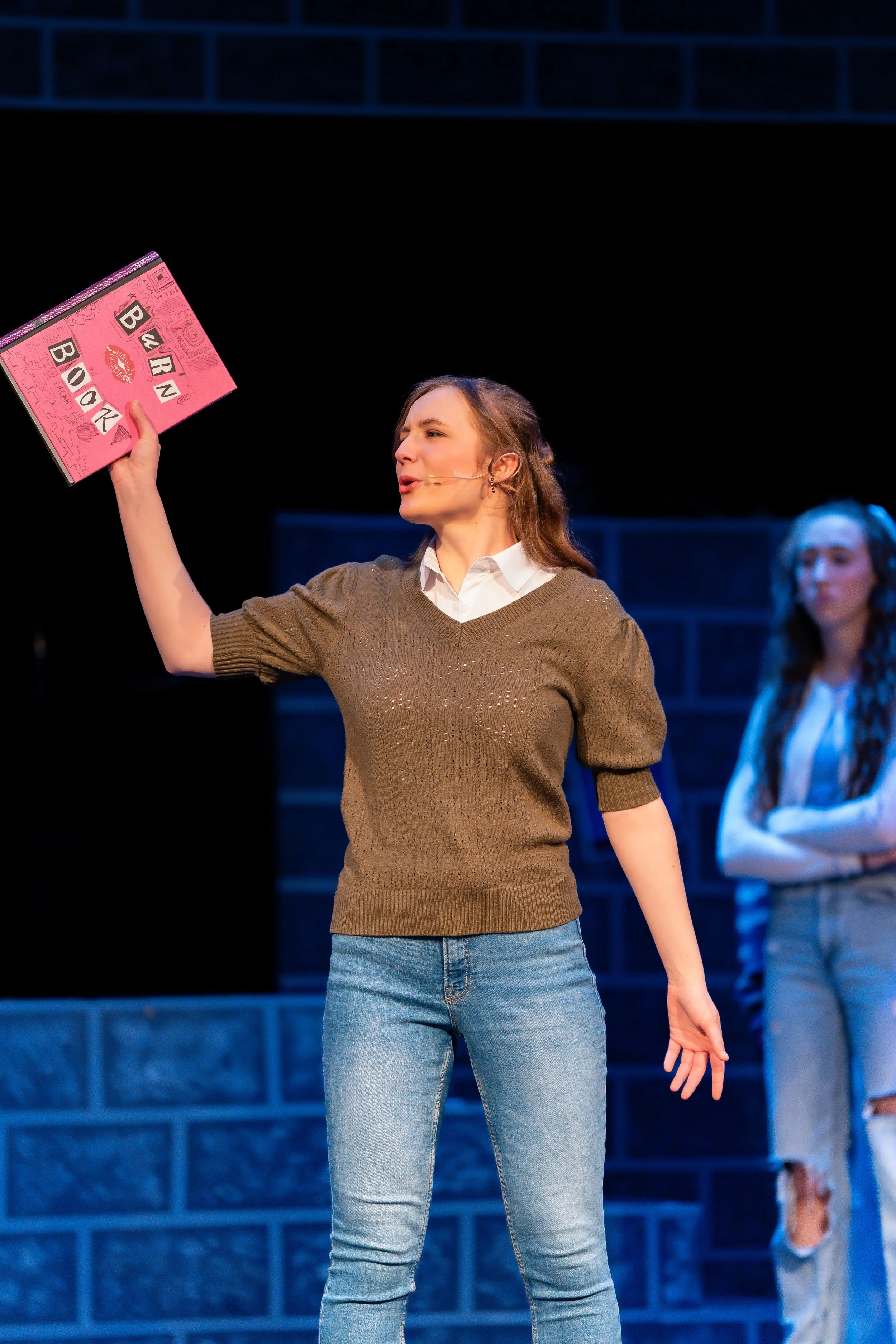A woman on stage holding a pink sign that reads 'BURN BOOK,' with another woman standing in the background.