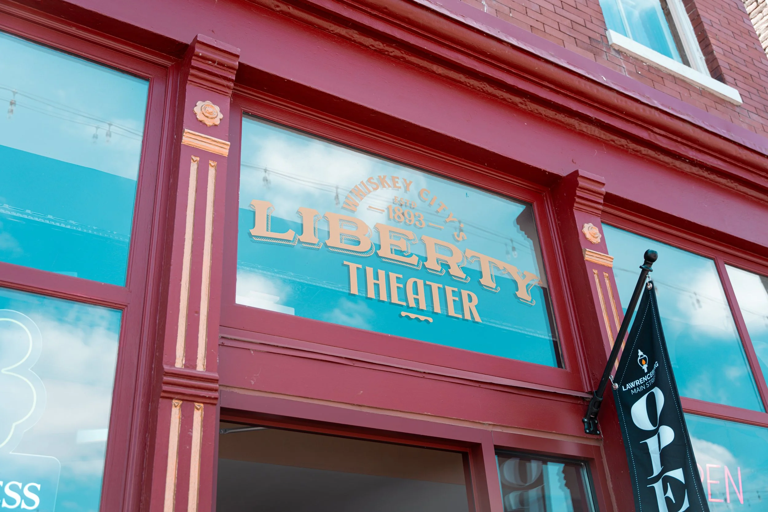 Red storefront with large window displaying the words "Liberty Theater" established 1893, and part of a black banner with white text hanging outside.