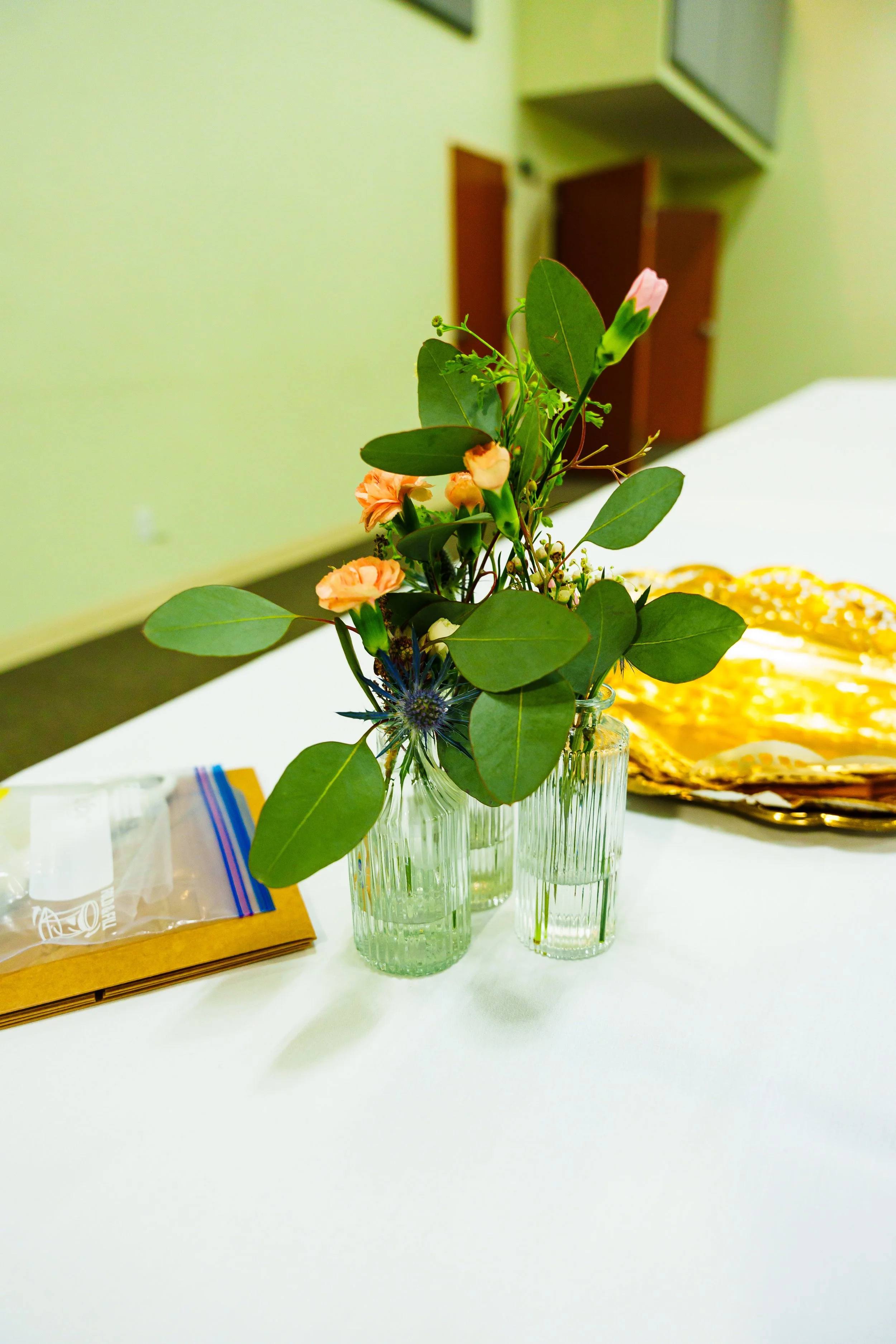 A small bouquet of pink and peach flowers with green leaves in three clear ribbed glass vases on a white table, with a yellow cloth and some plastic bags nearby in a room with pale green walls and wooden cabinets.