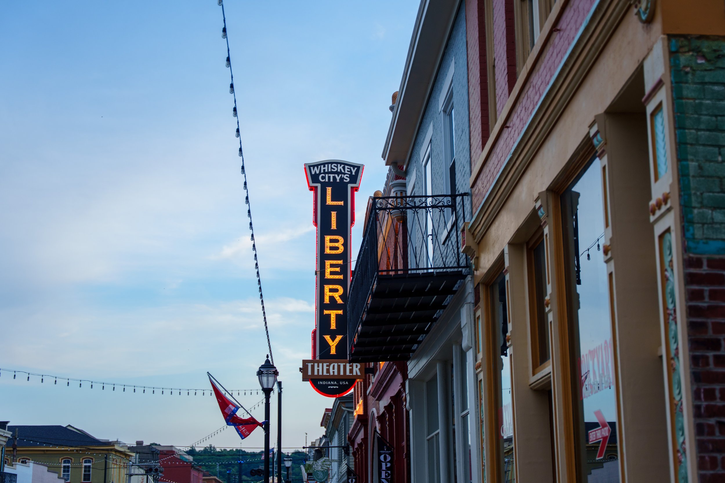 Street view of a downtown area with colorful buildings, string lights overhead, flags, and a neon sign for Whiskey City's Theater, located in Indiana, USA.