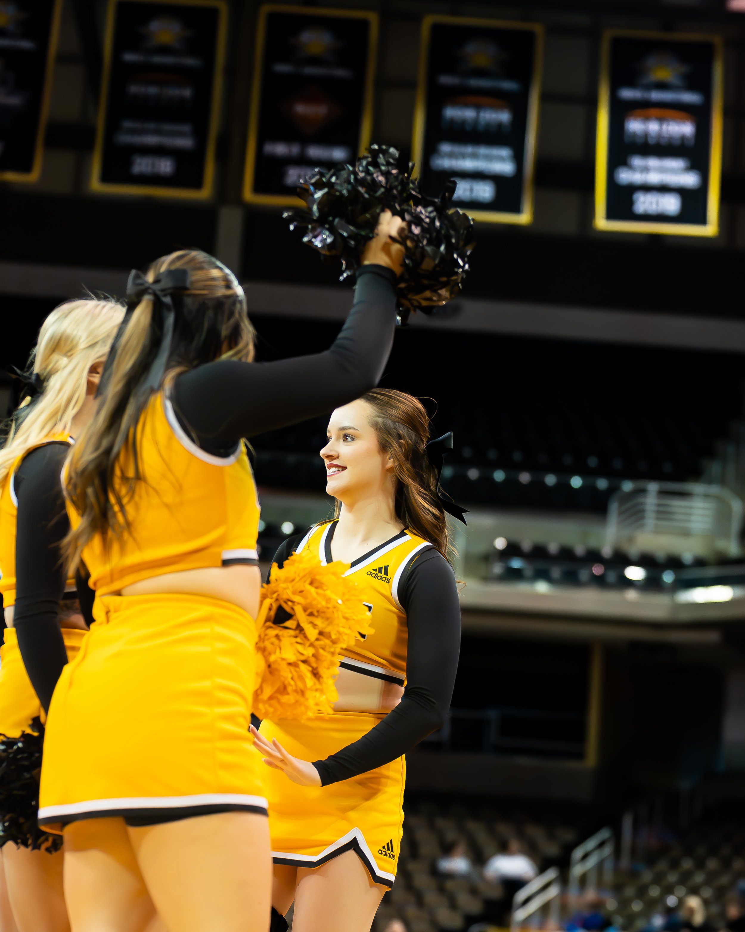 Cheerleaders in yellow and black uniforms with pom-poms, one cheerleader holding a pom-pom high, standing in an indoor arena.