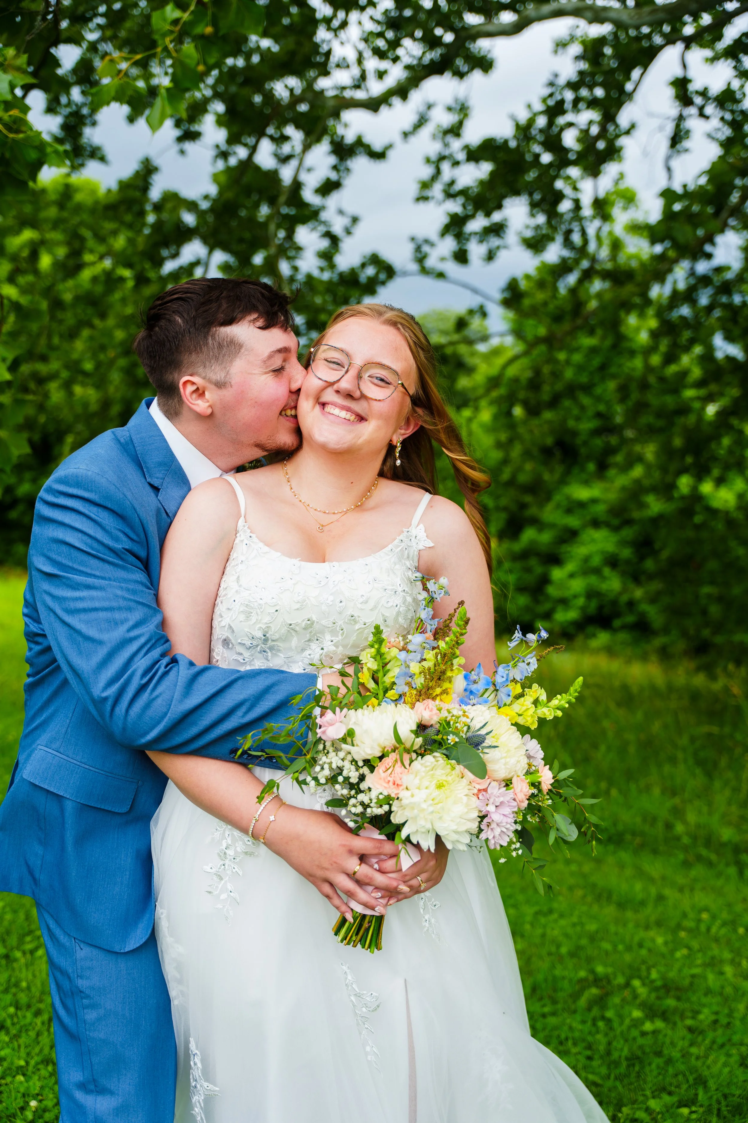 A young couple dressed in wedding attire outdoors. The groom is in a blue suit, kissing the bride’s cheek. The bride is smiling, holding a bouquet of white, pink, and purple flowers, wearing glasses and a white lace wedding dress, with long hair. Gre
