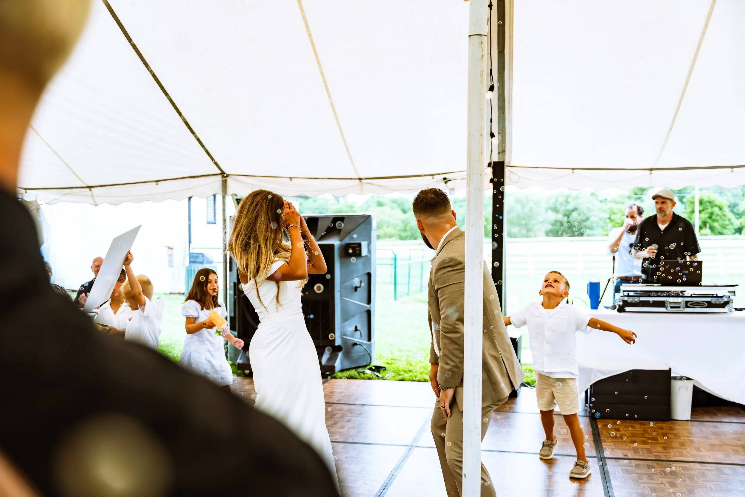 People dancing under a white canopy tent at an outdoor event, with a DJ setup in the background and children participating.