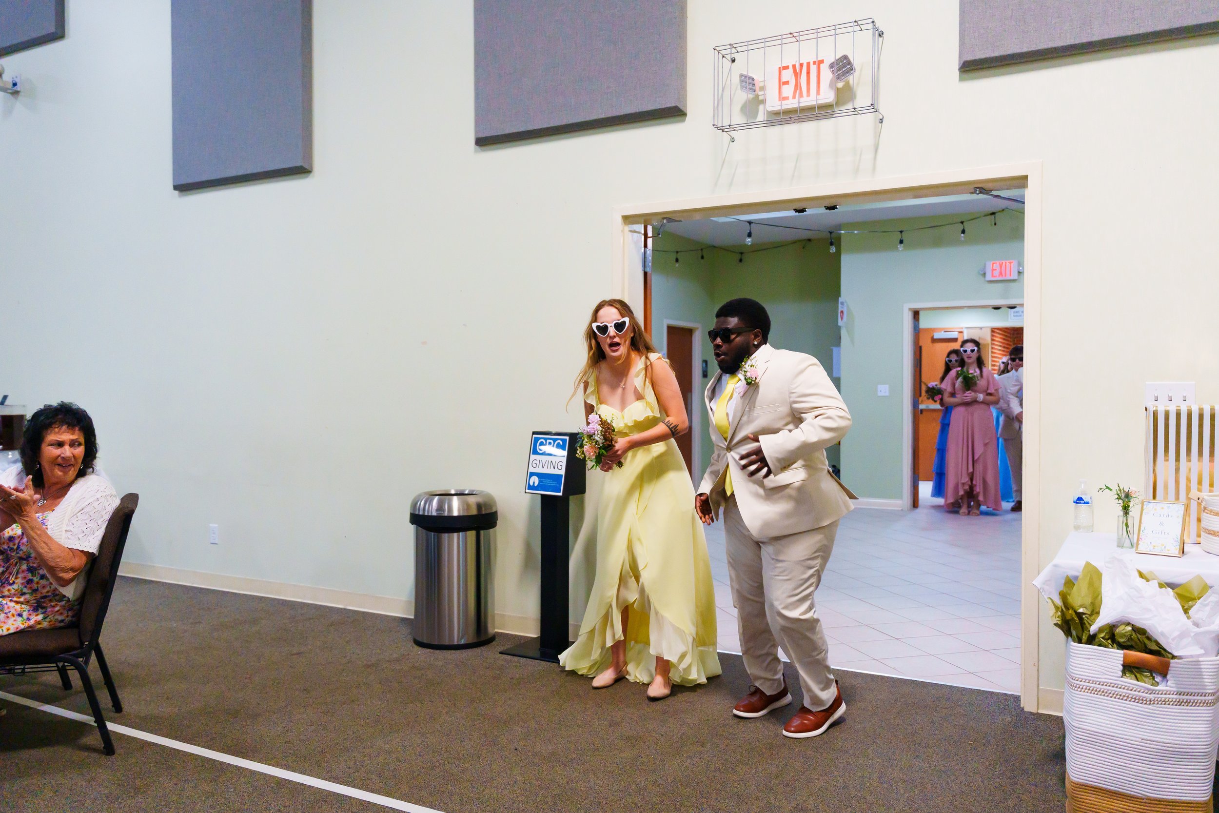 A wedding reception with guests, including a woman in a yellow dress with heart-shaped sunglasses and a man in a beige suit, both dancing. Other guests are seated and watching, with some holding bouquets. The setting features a sign for giving and ex
