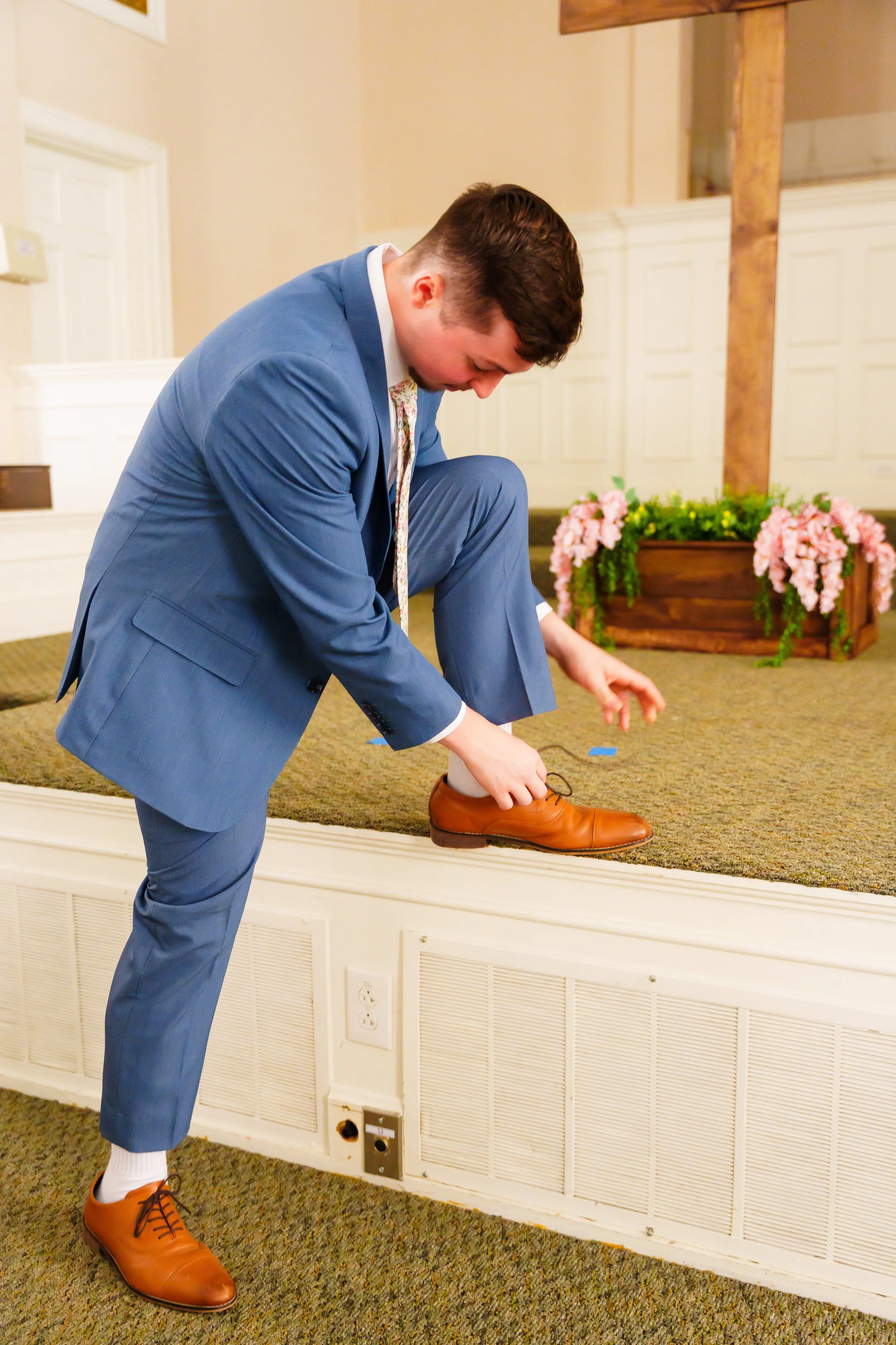 A man dressed in a blue suit tying his brown leather dress shoe on a carpeted stage with floral decorations in the background.