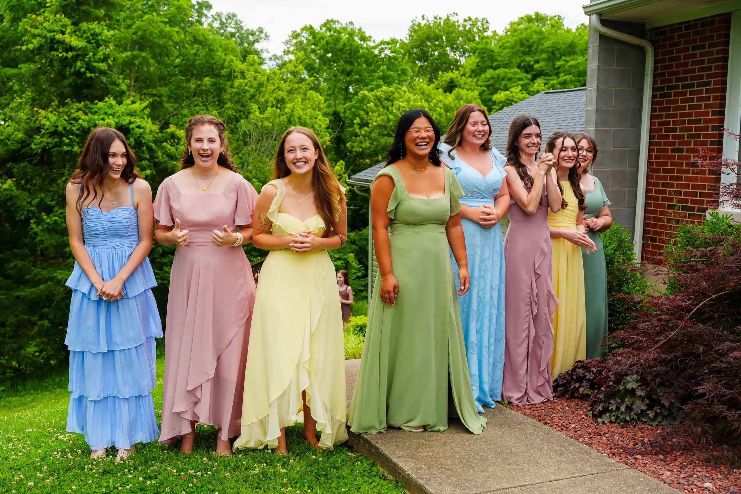 Group of women in colorful dresses standing outdoors on a lawn near a brick house, smiling and laughing together.