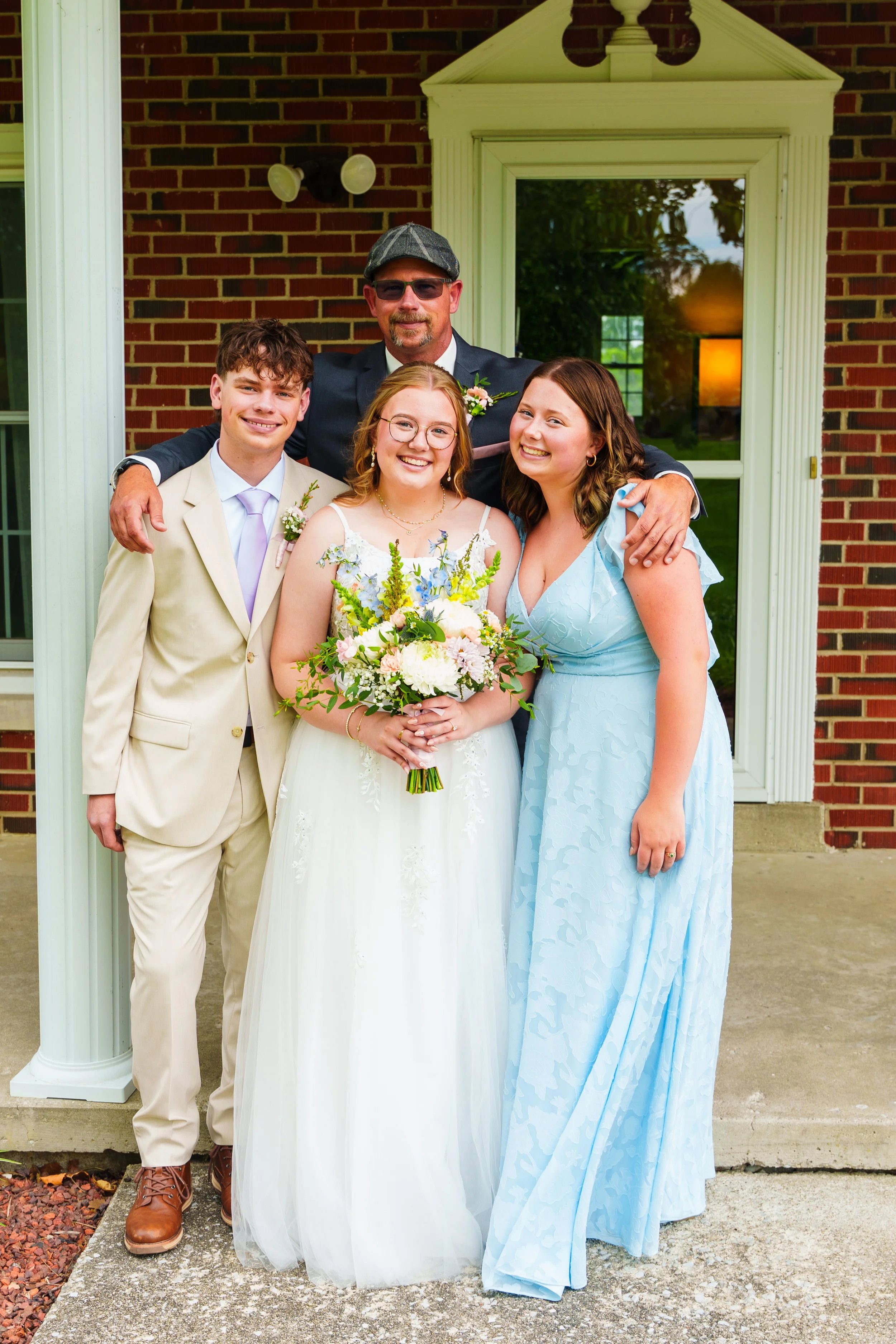 A group of four people, including a bride in a white wedding dress holding a bouquet, a groom in a beige suit, and two women in pastel dresses, standing outside a brick building during a wedding celebration.