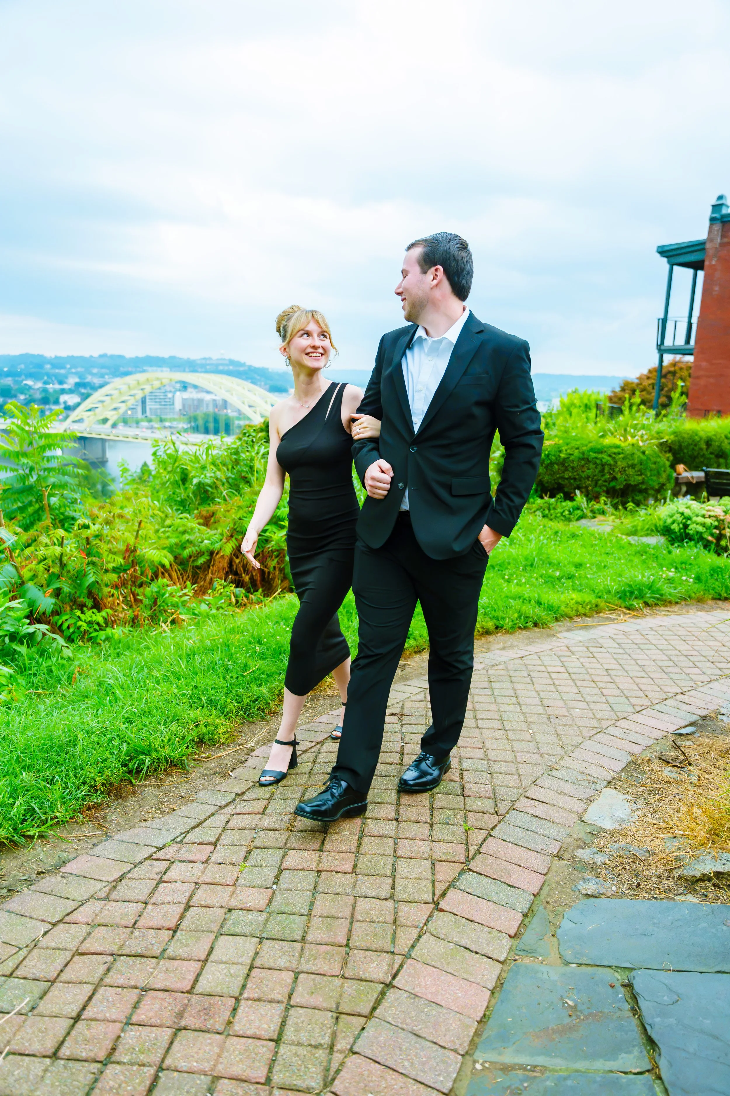 A man and woman dressed in formal attire walking arm-in-arm on a brick path in a park, with a city skyline and a distinctive bridge in the background.