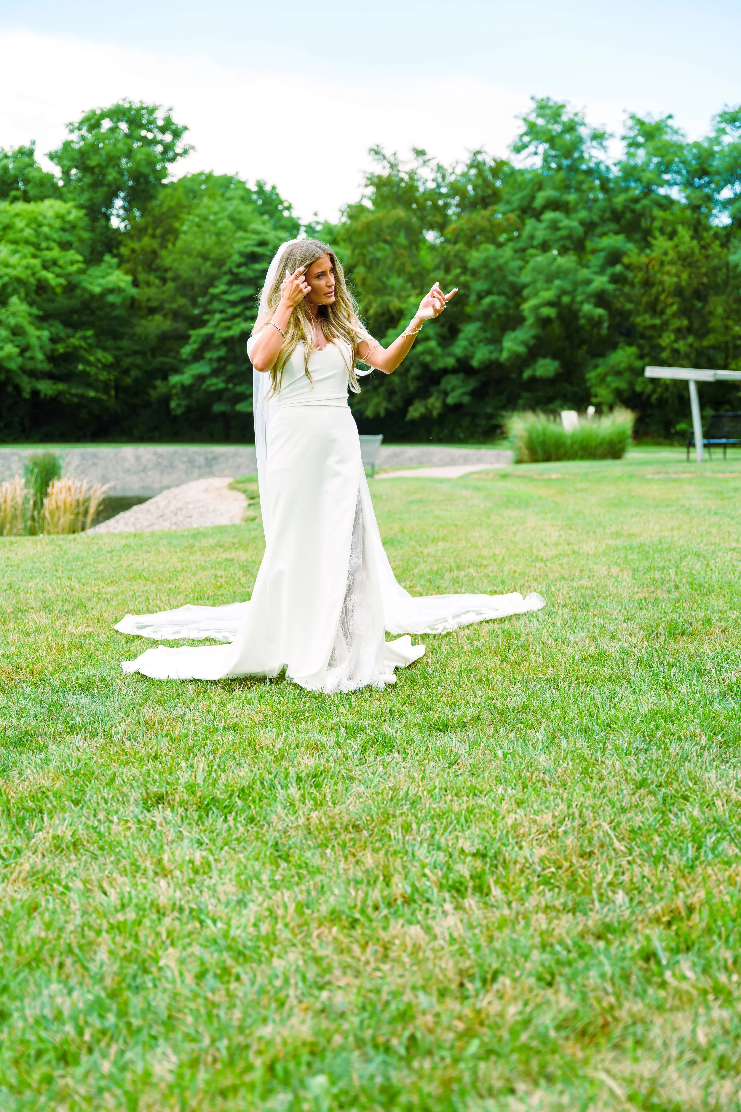 A woman wearing a white dress and veil, likely a bride, is standing on a grassy lawn in a park with green trees in the background, looking thoughtful and touching her hair.