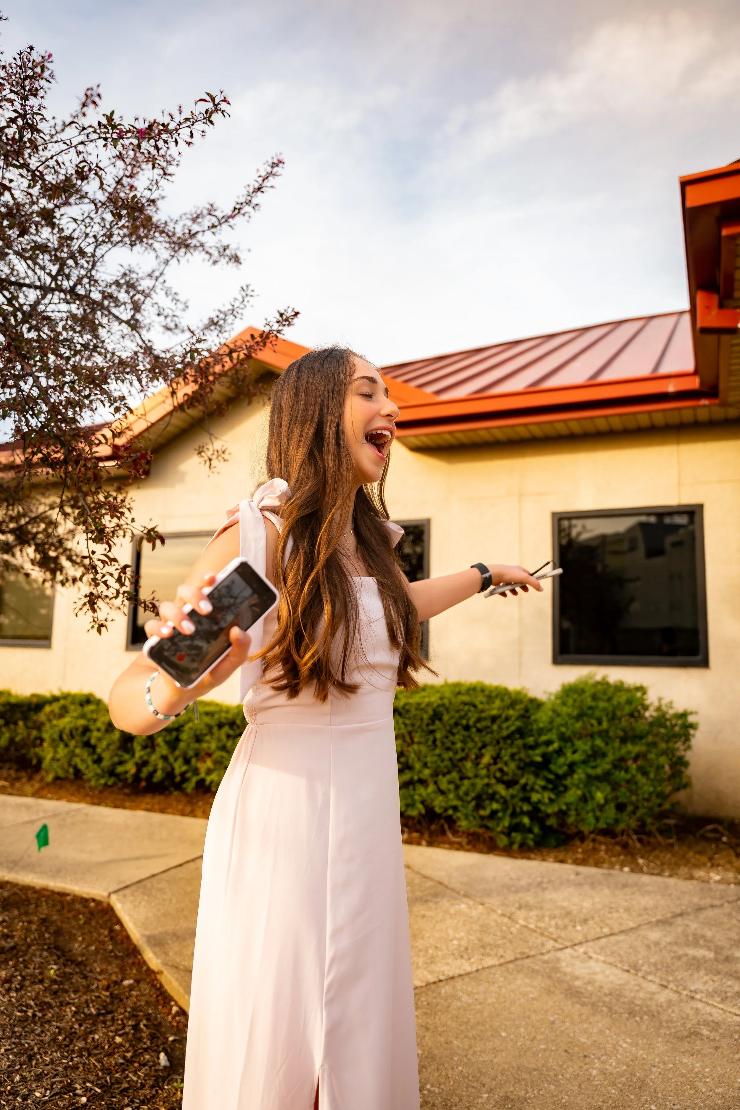 Young woman with long brown hair in a white dress smiling and talking outdoors during sunset, holding a smartphone.