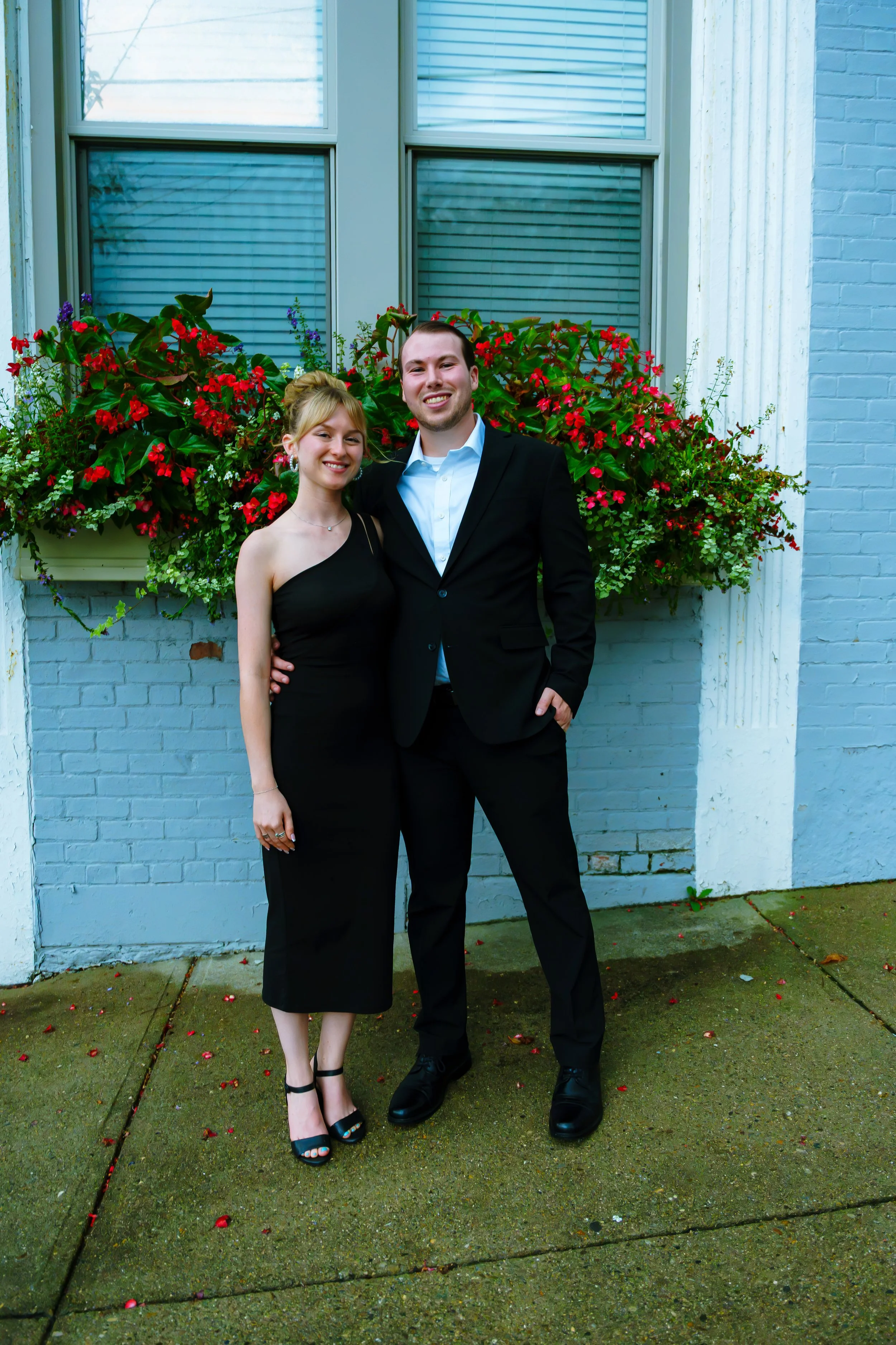 A smiling young couple dressed formally stands in front of a colorful flower garden against a light blue brick wall. The woman wears a black dress and heels; the man is in a black suit with a white shirt.
