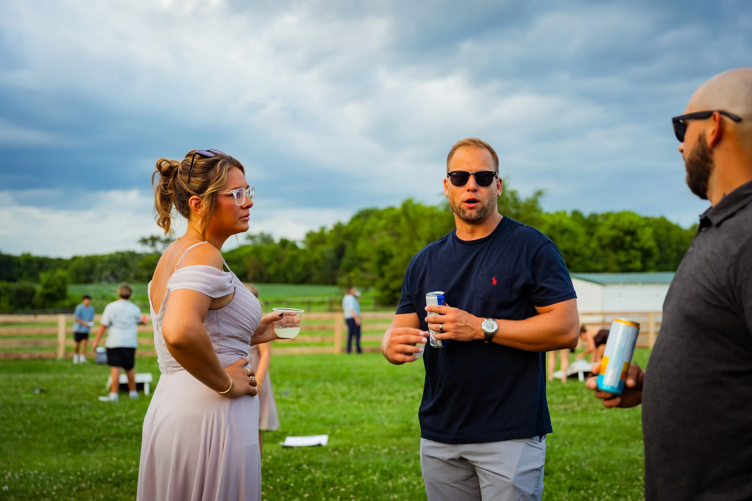 Three people engaged in conversation at an outdoor gathering on a grassy field. The woman on the left is wearing glasses and a light dress, holding a drink, while the man in the middle is wearing sunglasses, a dark t-shirt, and holding a drink. The t