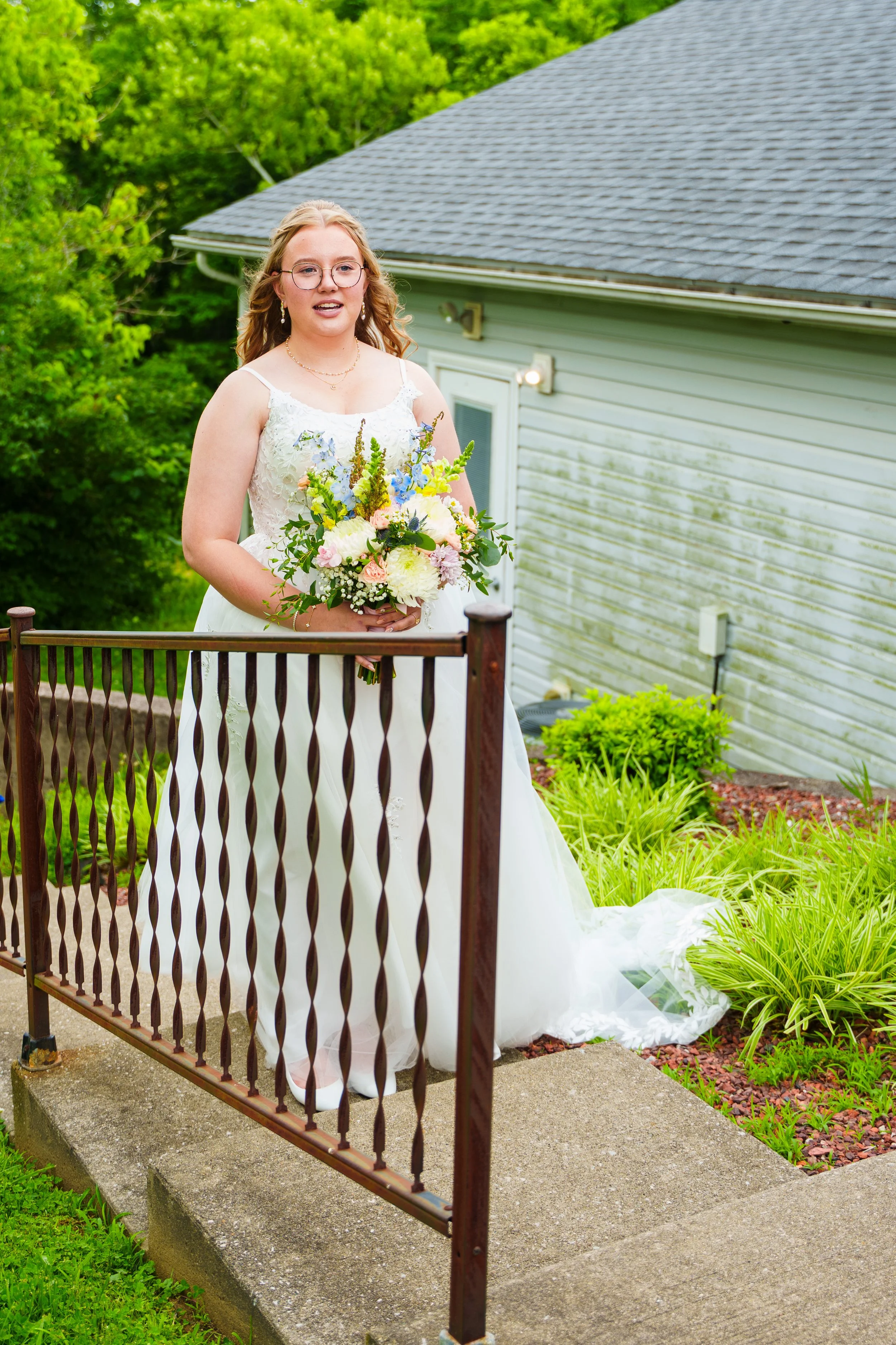 A bride in a white wedding dress holding a colorful bouquet standing behind a metal gate on a concrete step in a garden, with a light green house and green trees in the background.