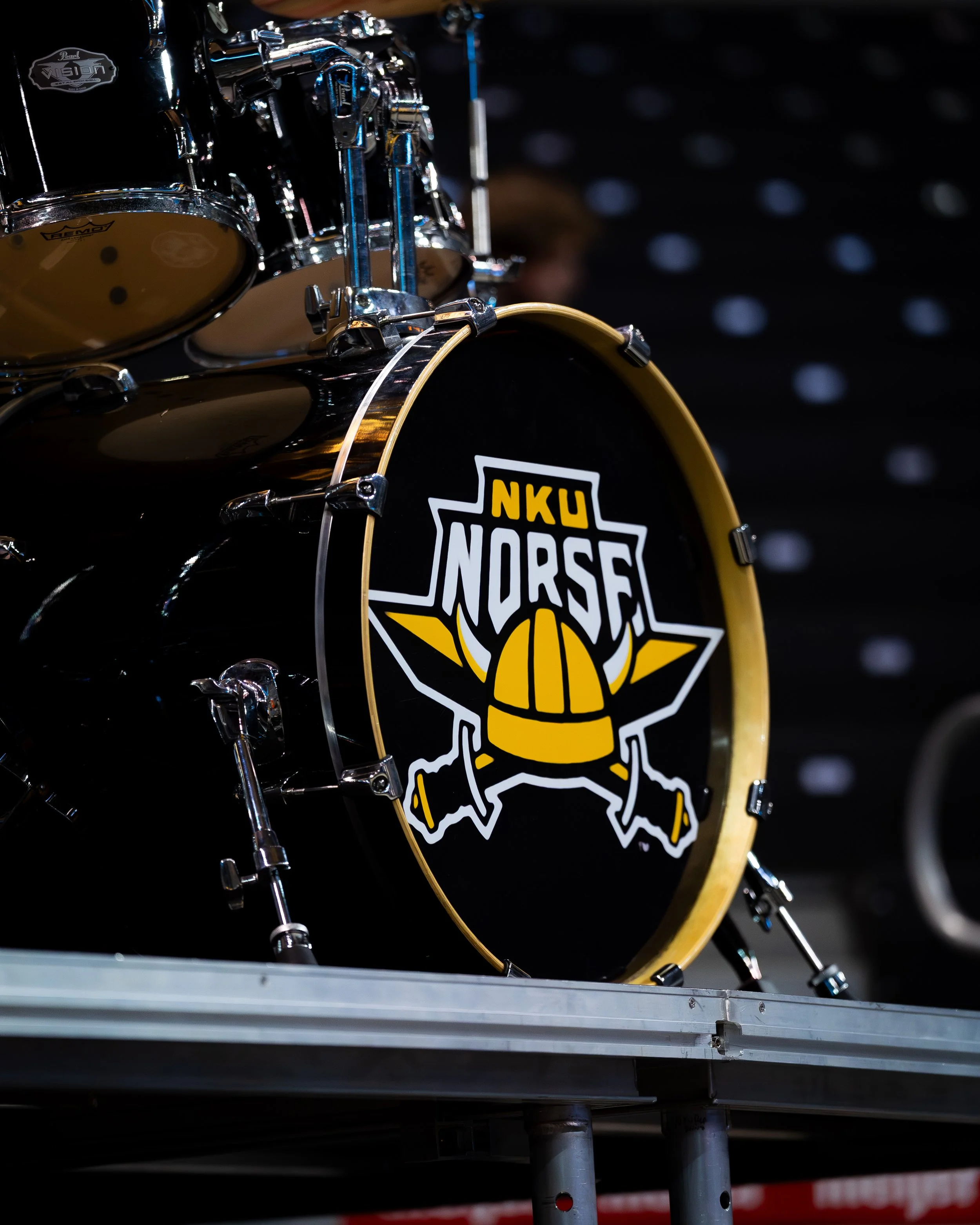 Close-up of a drum set on a stage with a sticker that reads 'NKU NORSE'. The drum set features shiny metallic hardware and a black drum with a yellow rim. The background is dark with out-of-focus lights.