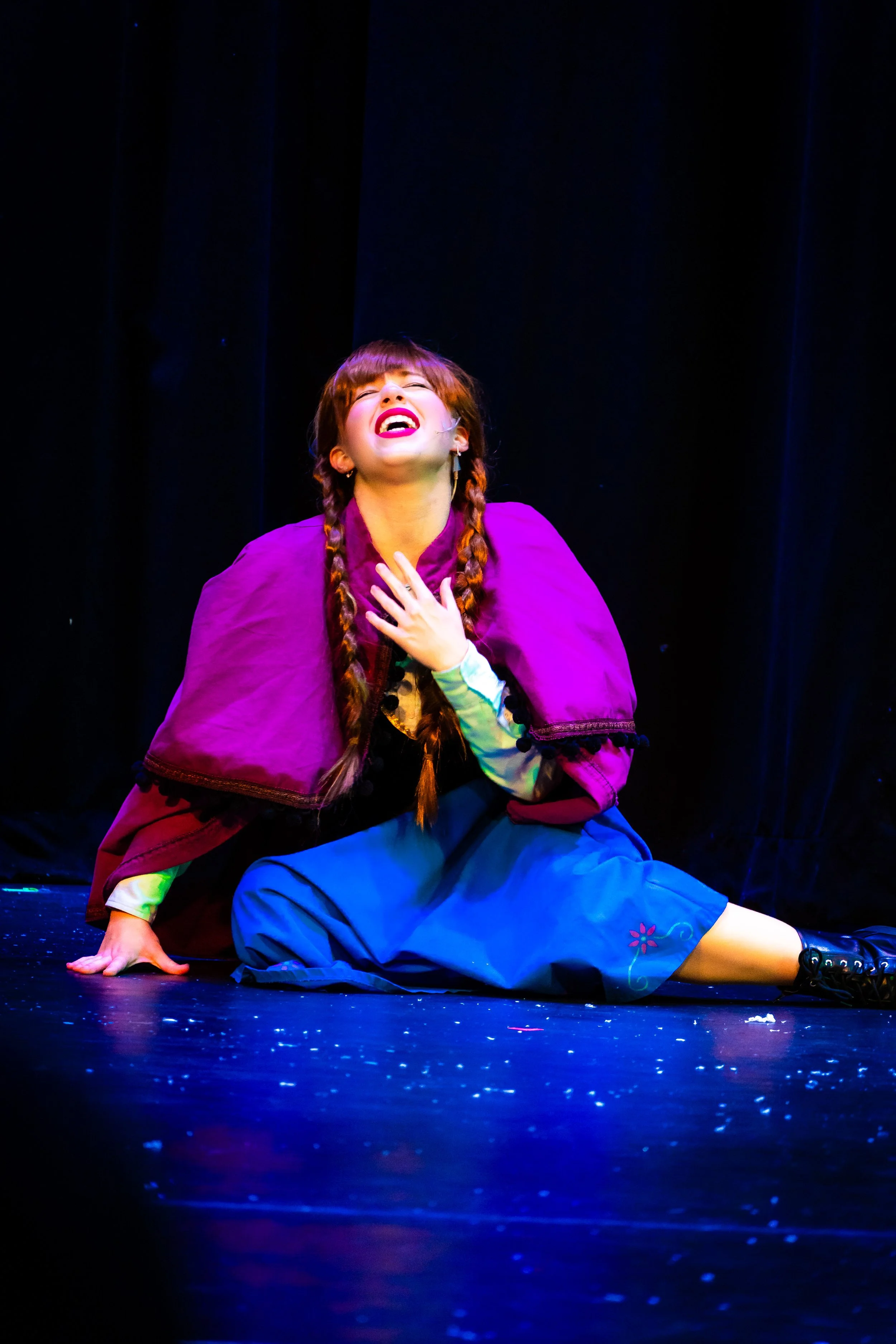 A woman with braided red hair, wearing a purple cape, green dress, and blue skirt, is sitting on the stage floor, holding her chest and laughing with her eyes closed.