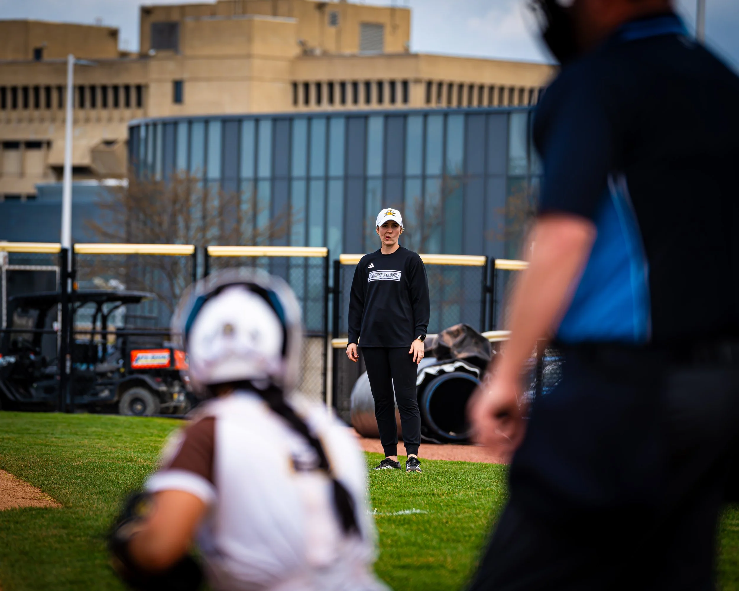 A baseball coach or instructor speaking to players on a baseball field, with some standing and some sitting, during a practice or game, with a large building in the background.