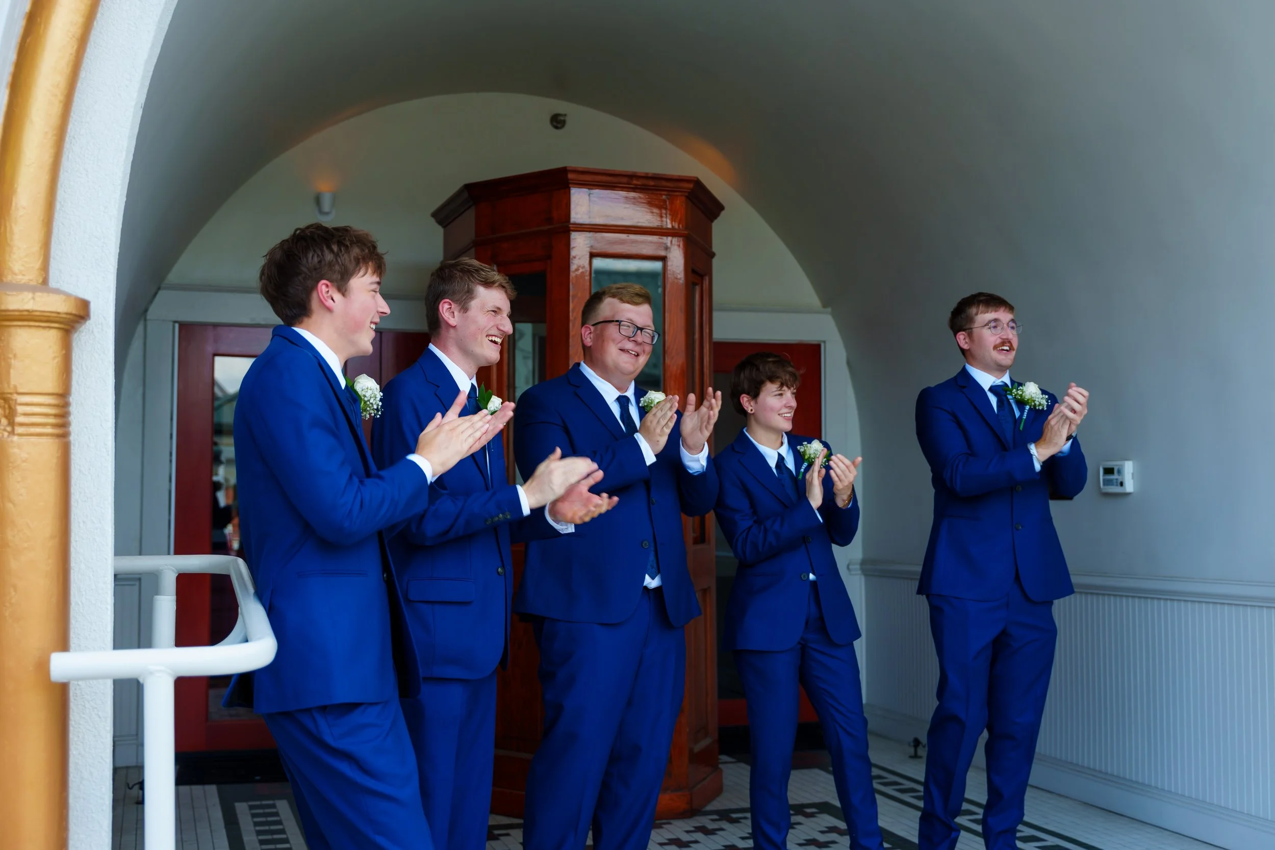 Five young men dressed in matching blue suits and white shirts standing indoors, clapping and smiling at what appears to be a wedding celebration.