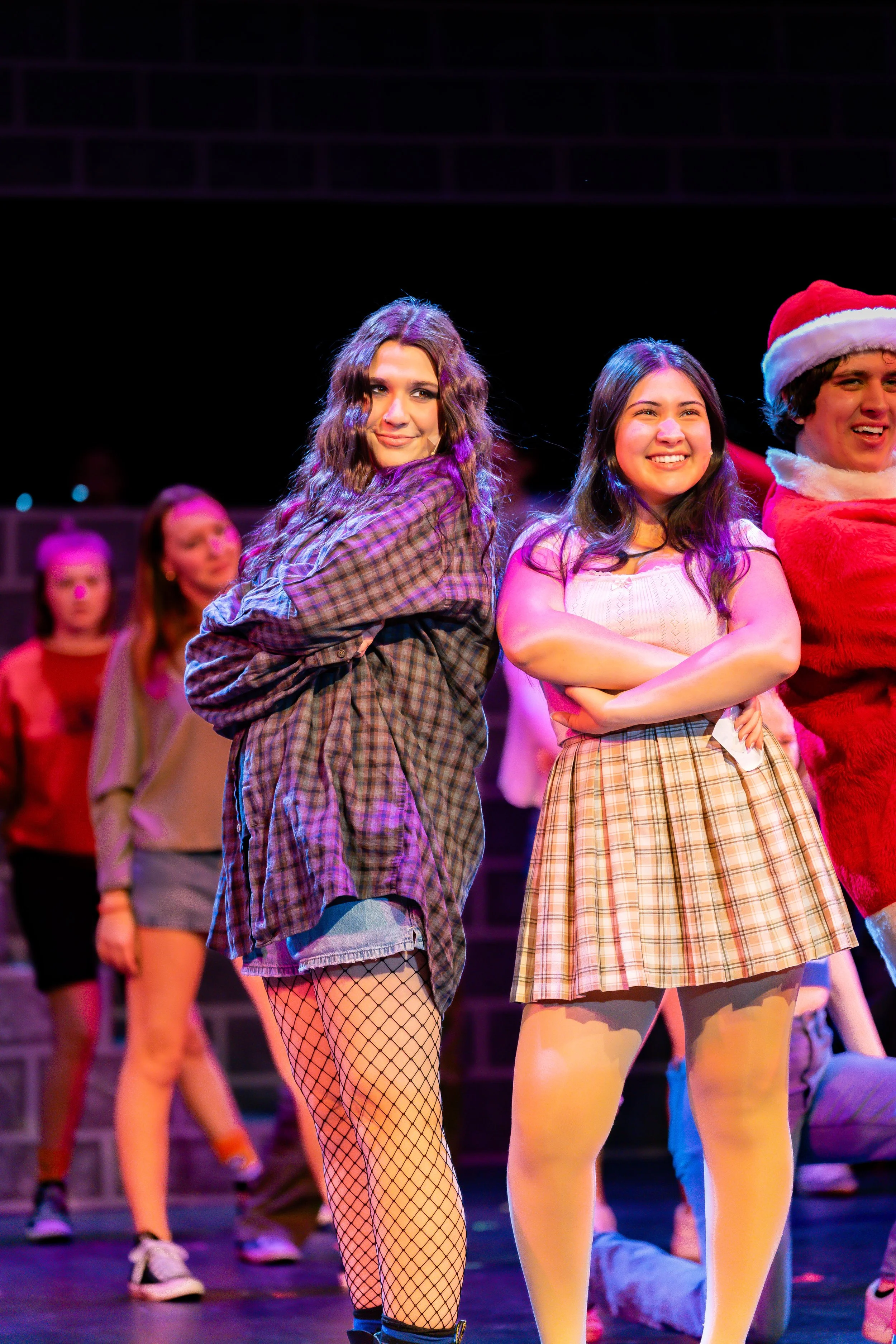 People performing on stage in costumes, including two women in plaid and beige skirts with fishnet and tights, and a man in a Santa suit with a Santa hat, during a holiday-themed event.