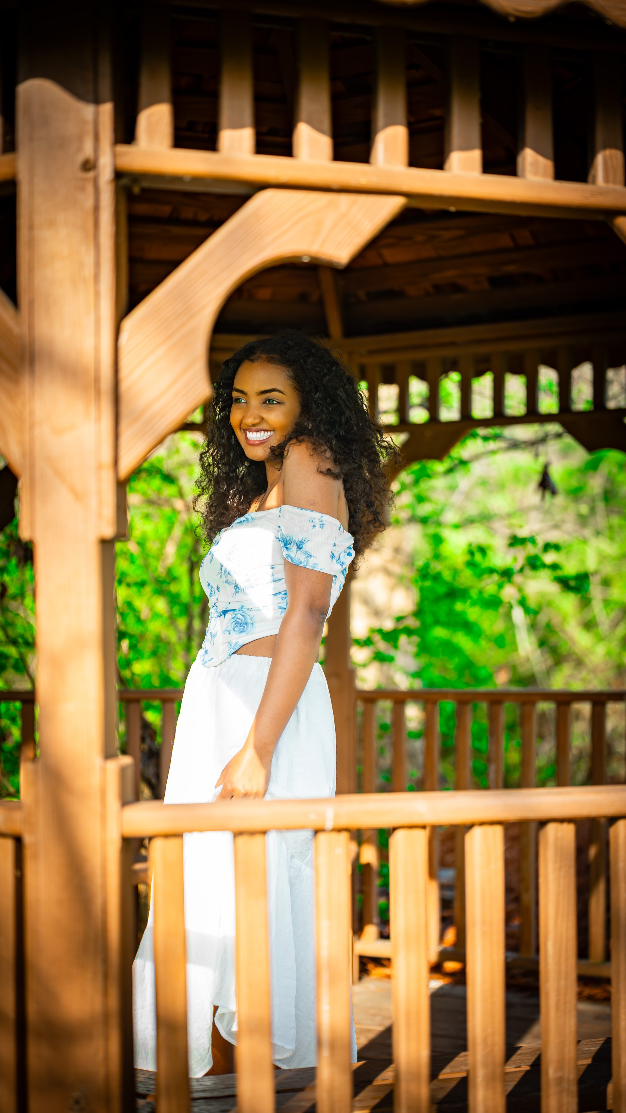 Smiling woman with curly hair standing on a wooden balcony or deck surrounded by green trees.