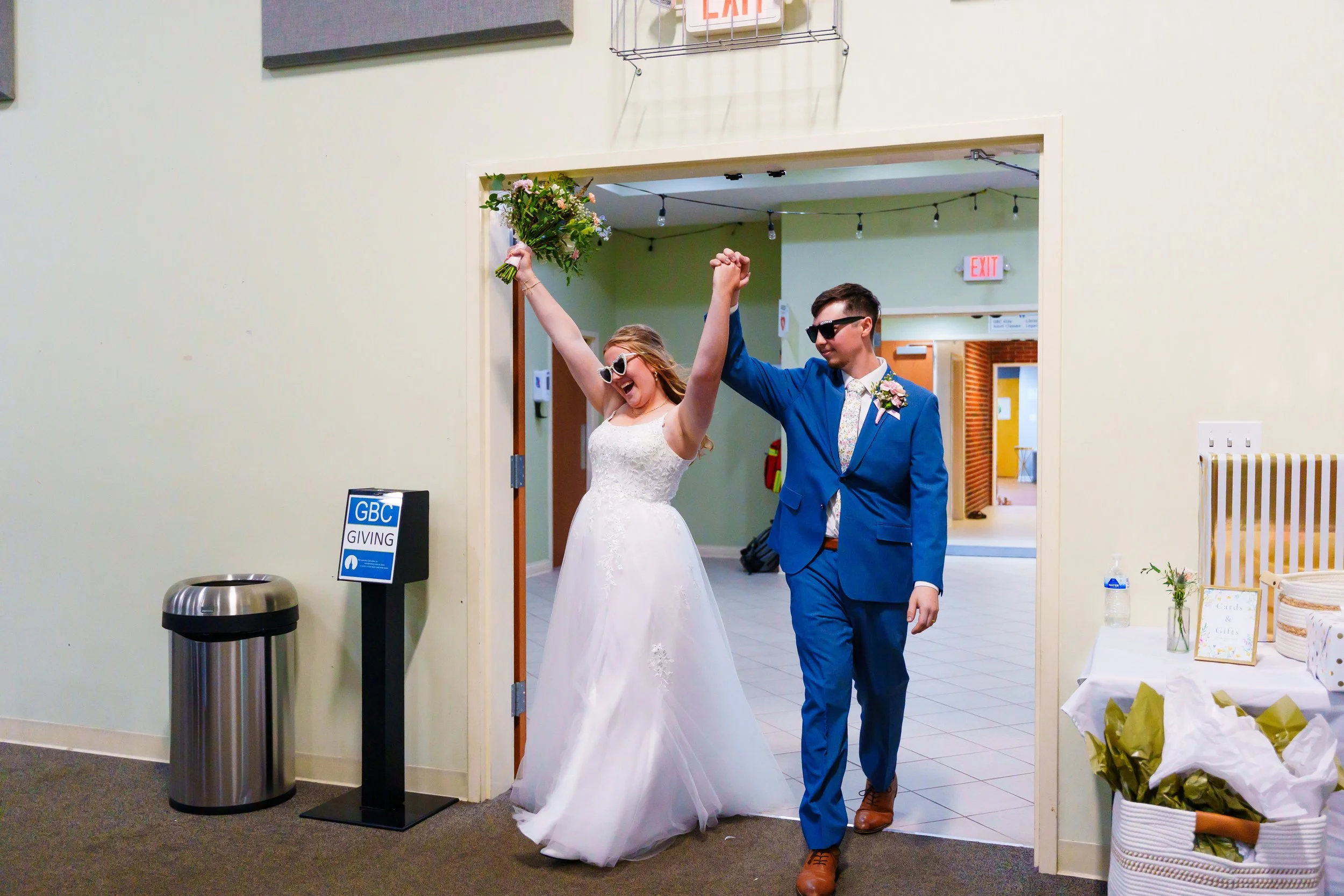 A bride in a wedding dress holding a bouquet and smiling as she walks with a man in a blue suit, sunglasses, and a tie, inside a building decorated for a celebration, possibly a wedding reception.