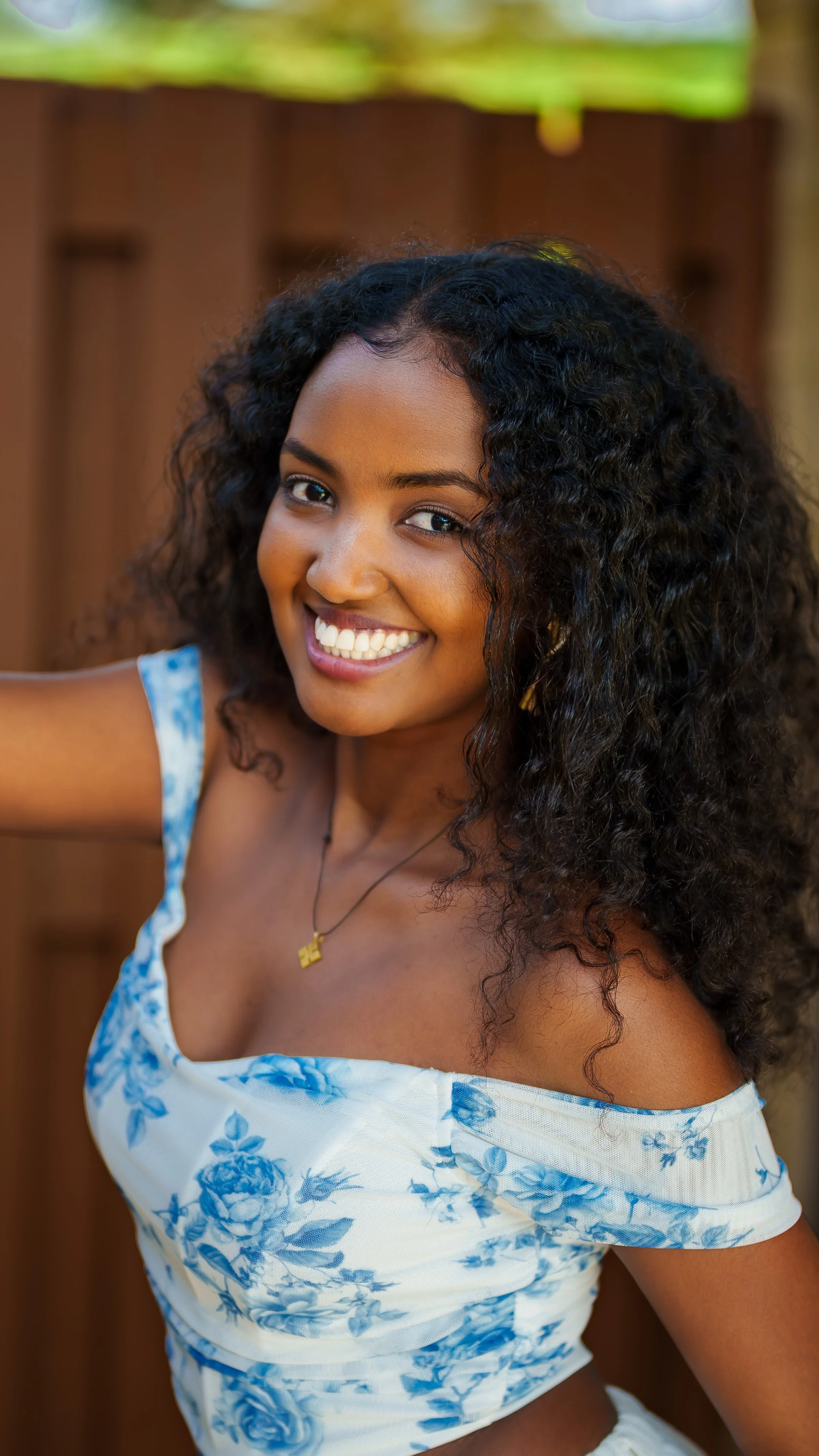 A young woman with curly black hair smiling at the camera, wearing a white off-shoulder top with blue floral pattern and a gold necklace.