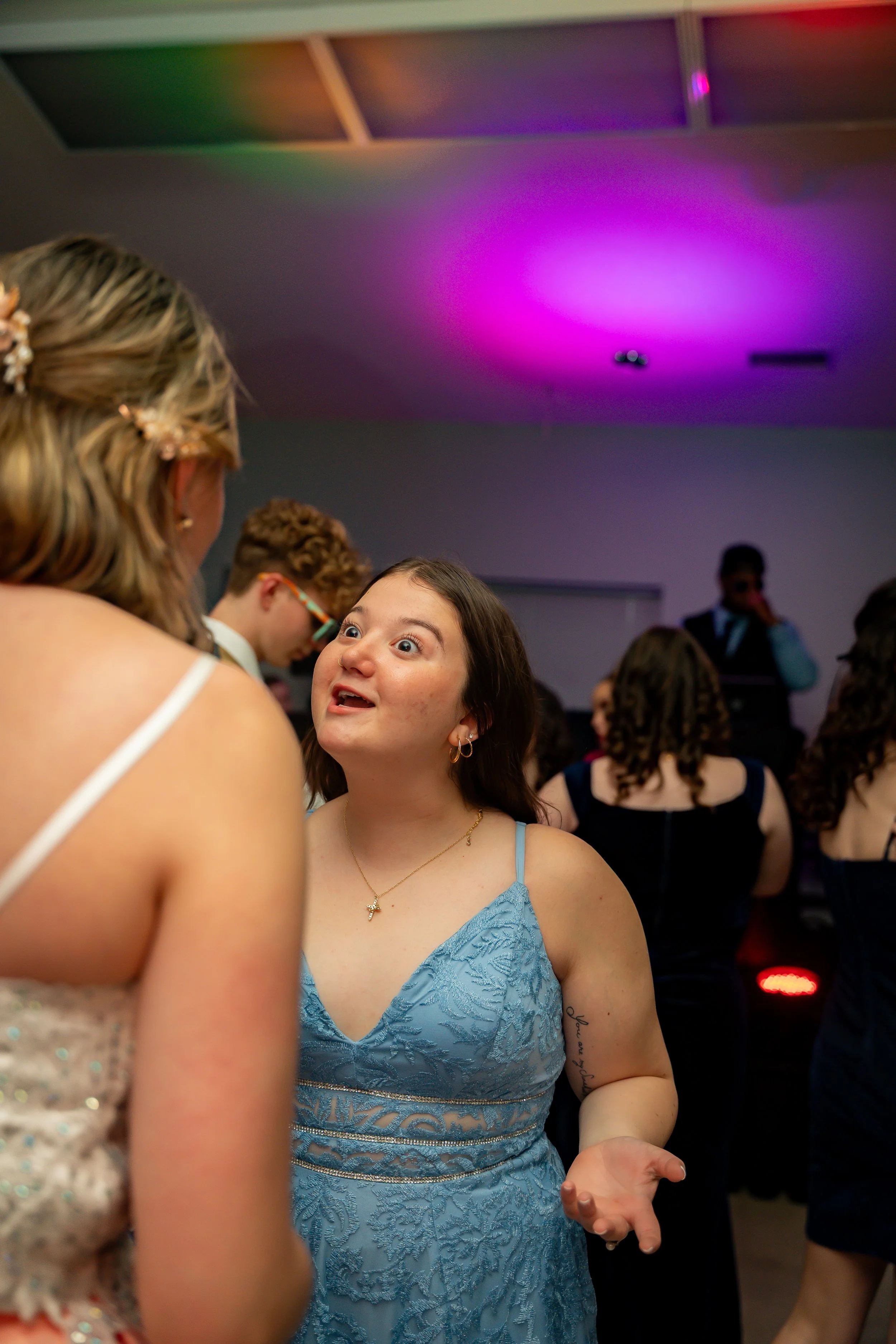 A young woman with dark hair and earrings, wearing a blue lace dress, at a party or event, talking to someone. People are in the background, and the ceiling has purple lighting.