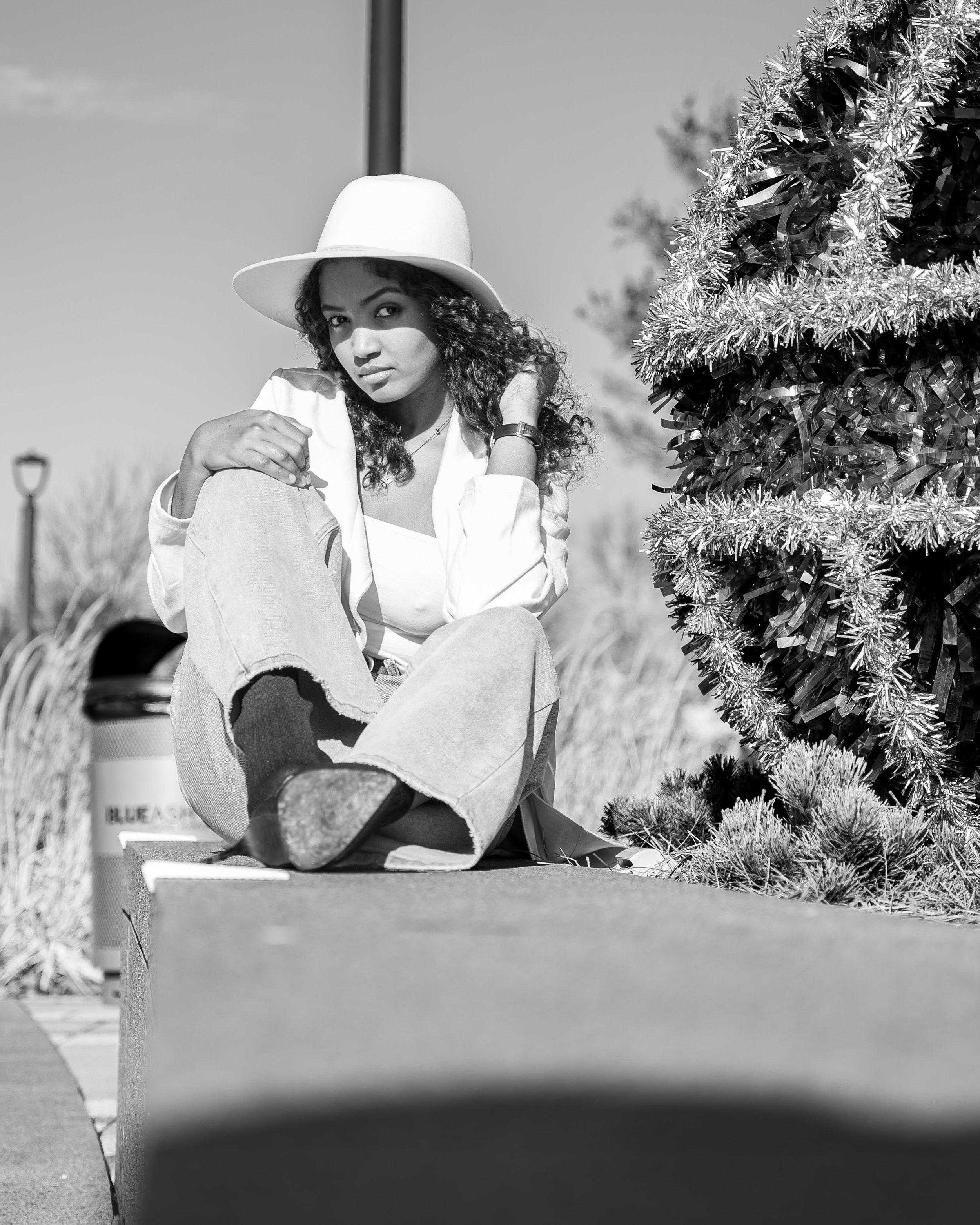 A woman wearing a wide-brimmed hat, a light-colored blazer, and jeans, sitting outdoors next to a decorated Christmas tree, with a trash can and lamp post in the background.