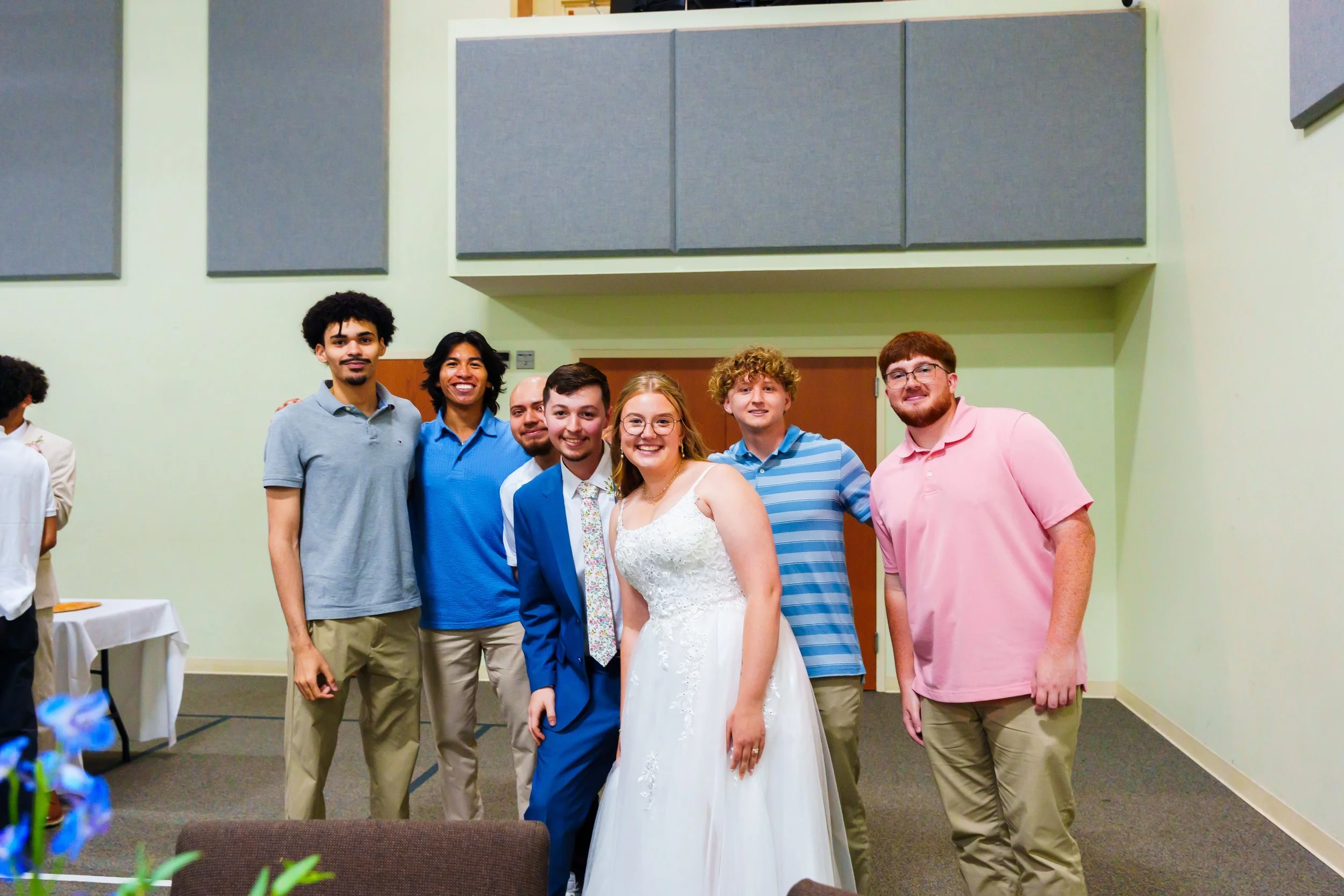 Group of seven young adults, including a bride in a white wedding dress, smiling and posing for a photo indoors against a light green wall with gray acoustic panels.