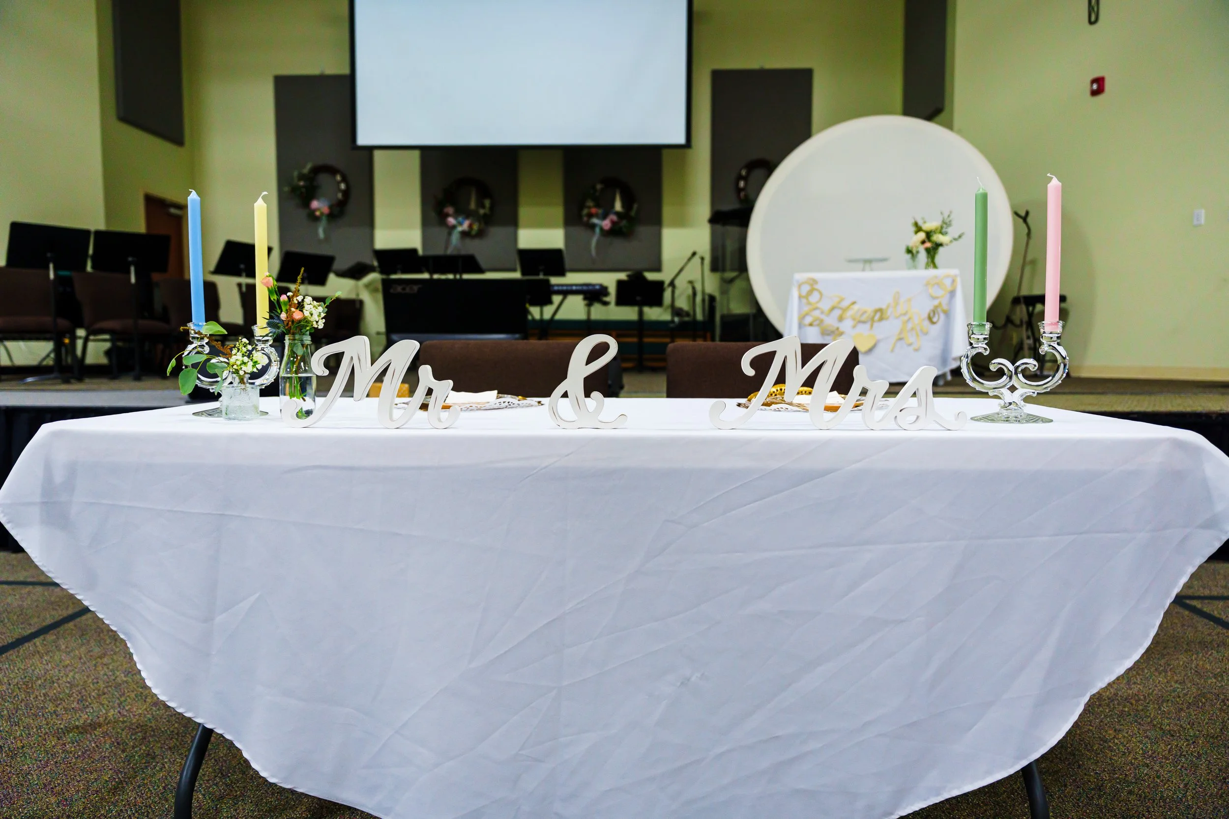 Wedding reception table decorated with pastel-colored candles, floral arrangements, and wooden signs reading 'Mr. & Mrs.' in a church hall with musical instruments and wreaths in the background.