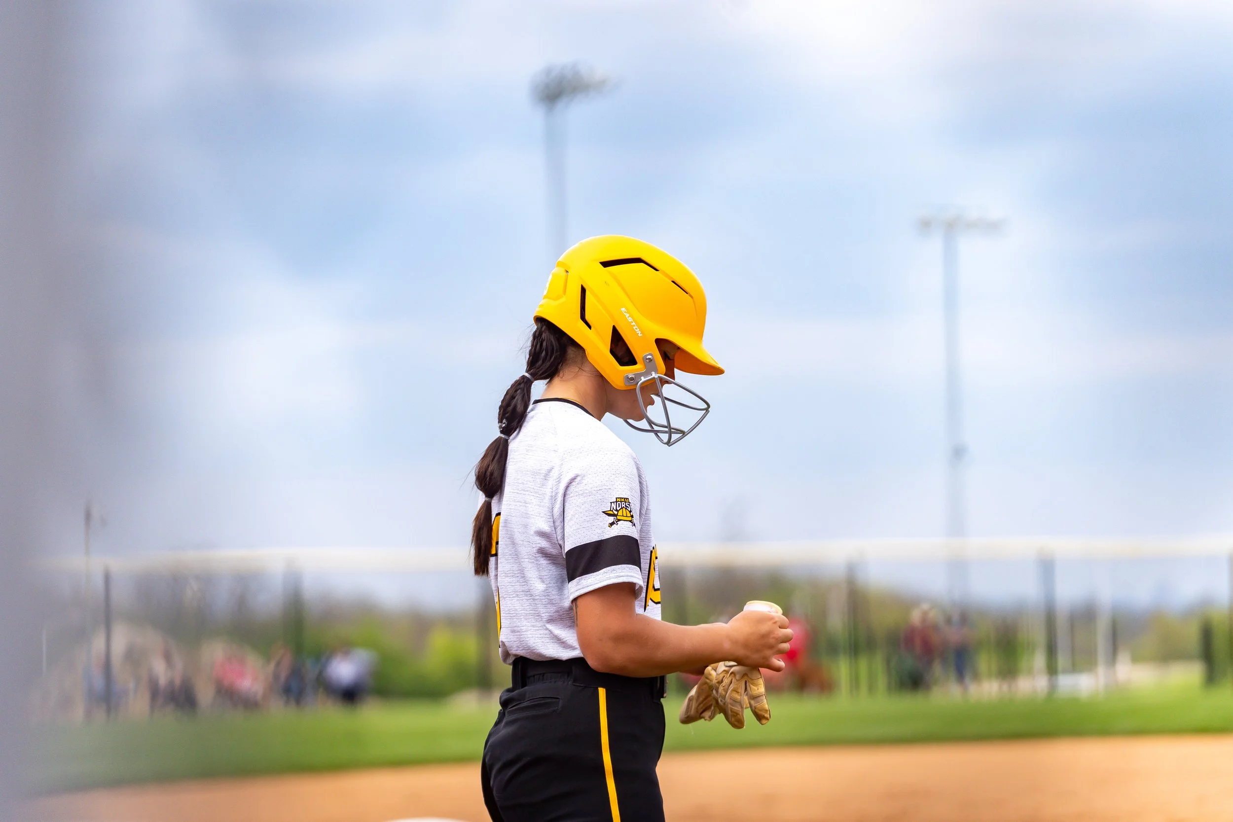 A female softball player wearing a yellow helmet with a face guard, a gray and black uniform, and gloves, standing on the field.