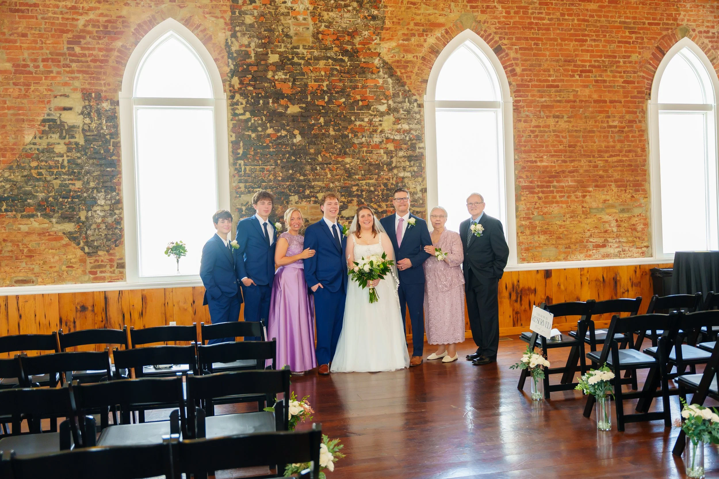 Group of people, including bride and groom, standing inside a church with brick walls and large windows, during wedding ceremony.