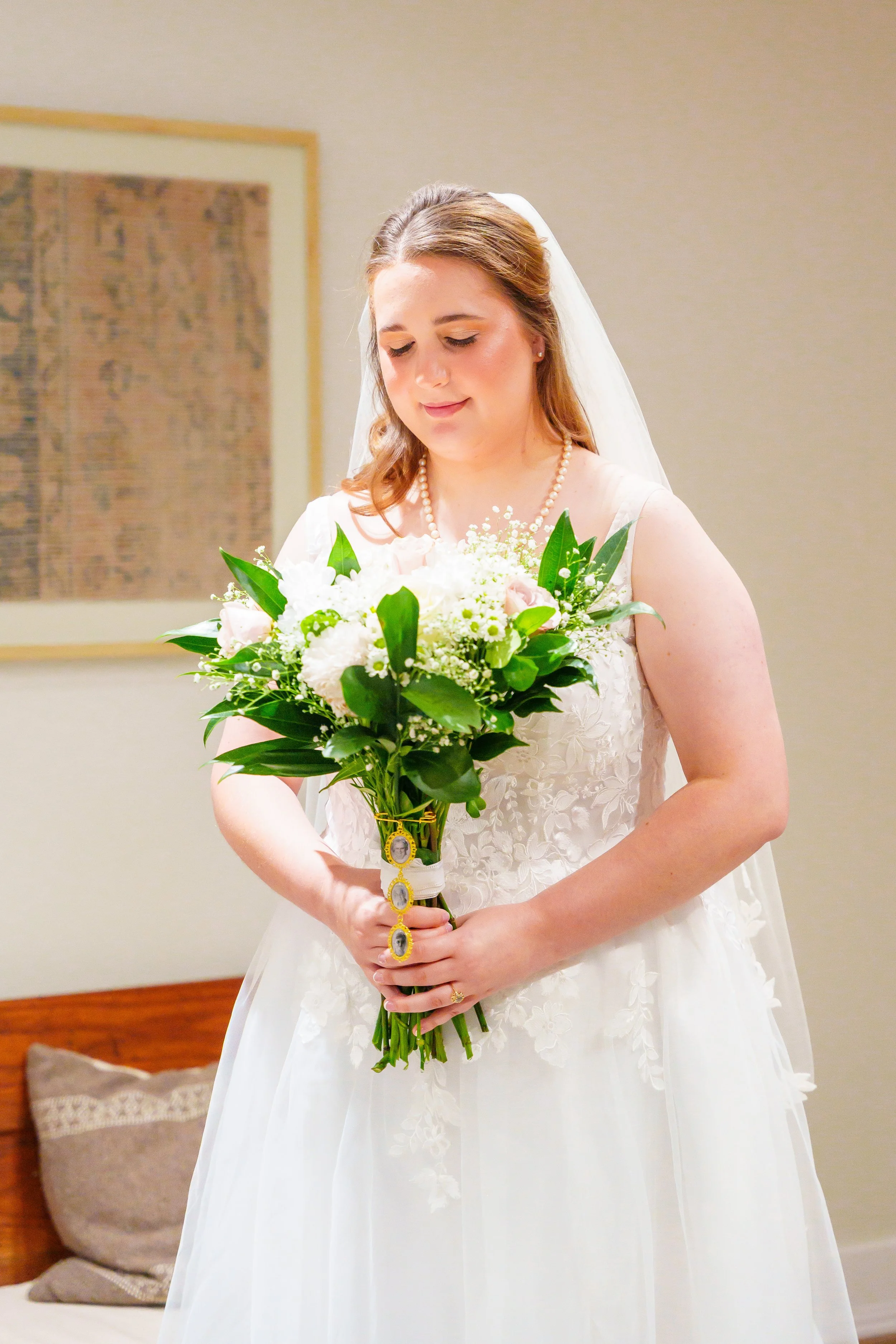 A bride wearing a white wedding dress with floral embroidery, a pearl necklace, and a veil, holding a bouquet of white and pink flowers with green leaves, smiling gently with her eyes closed.